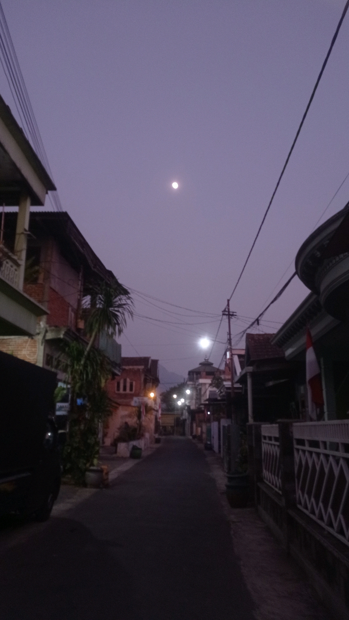 The image depicts a street scene in Mulyoagung, Indonesia, during dusk or pre-dawn. The sky is a muted purple-gray with a visible moon near the center. The street is lined with residential buildings, some appearing to be multi-story with visible windows and balconies. Utility poles and wires are prominent throughout the scene. A black vehicle is parked on the left side of the street, partially obscuring the view of a building and some greenery. There are streetlights illuminating parts of the road and the buildings. A fence with a diamond pattern is visible on the right side of the street. A flag, possibly red and white, is attached to a pole near a building on the right. The overall impression is a quiet, residential street in the early morning or evening hours.