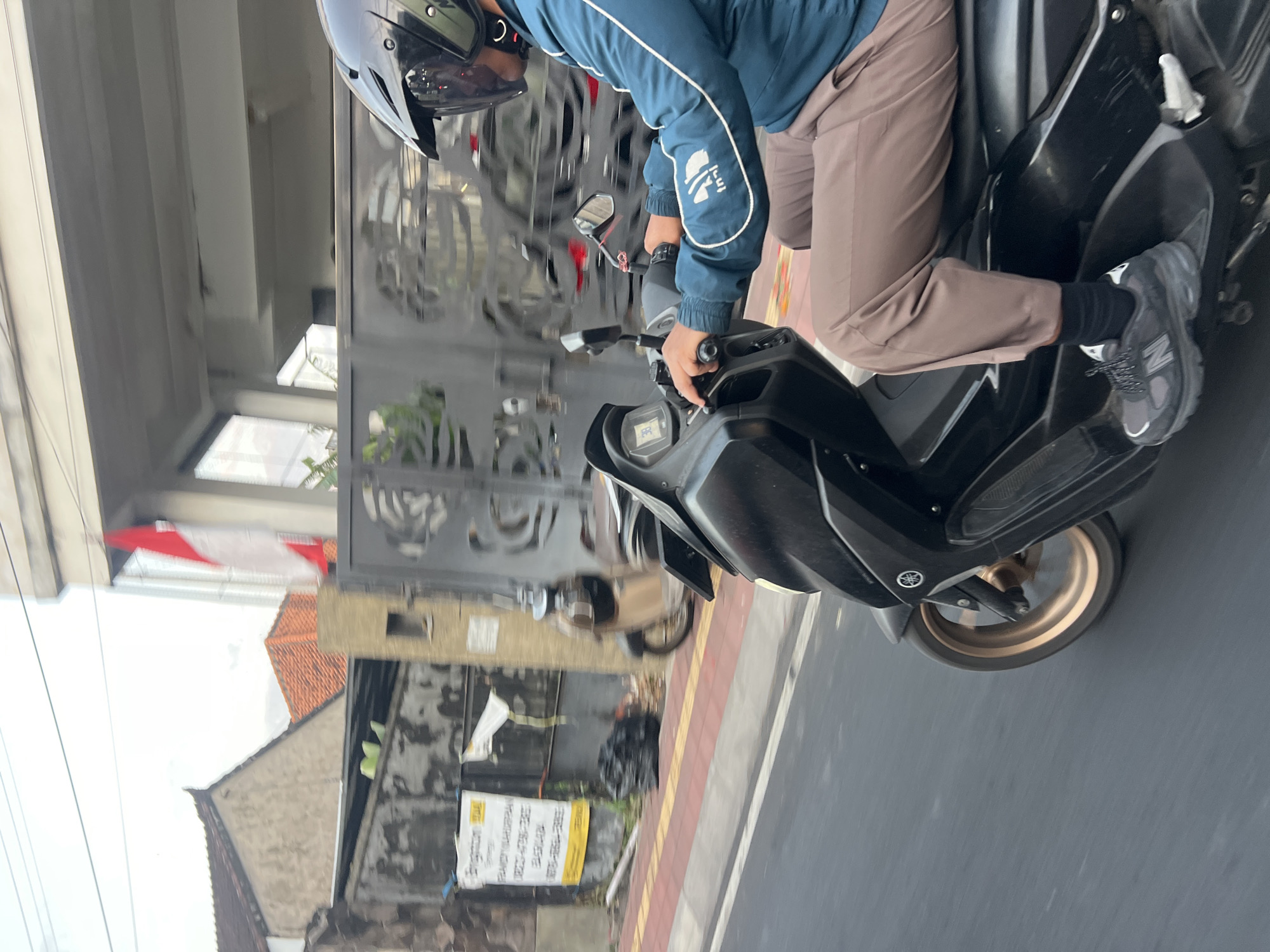 A person wearing a blue jacket, brown pants, and a helmet is riding a black scooter on a street in Denpasar, Indonesia. The scooter has a digital display showing "30". A red and white Indonesian flag is visible on a building in the background. The street has a red and yellow tiled sidewalk and a gray asphalt road. There is a black trash bag on the sidewalk. A metal gate with an ornate design is also in the background.