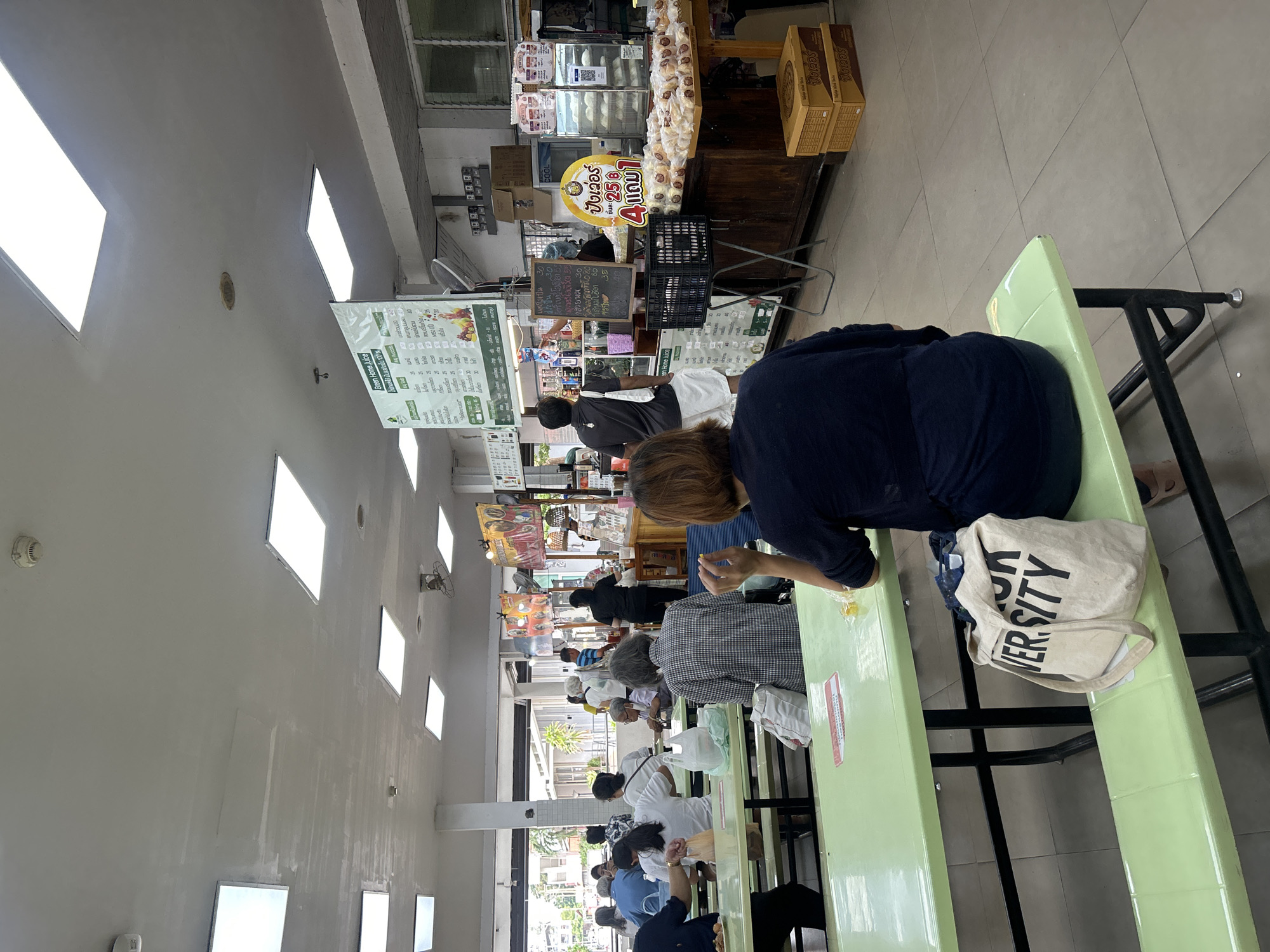 The image depicts a busy indoor dining area, likely a canteen or food court, in Tha Khlong Town Municipality, Thailand. The environment is characterized by long, light green picnic tables with attached benches, set on a tiled floor. Overhead, a white ceiling with fluorescent lighting panels is visible, along with air vents and a smoke detector.
Several individuals are seated at the tables, engaged in eating or conversing. In the foreground, a person is seated with their back to the camera, a canvas tote bag with the word "UNIVERSITY" printed on it rests on the bench beside them. Other patrons are scattered throughout the space, some with food items on their tables, while others are in the process of ordering or queuing.
At the rear of the dining area, a food stall or counter is operational, with staff members serving customers. Display shelves are stocked with various food items, and signage in Thai is prominent, including a large yellow sign with red text indicating a promotion. The overall atmosphere suggests a casual and functional dining establishment catering to a local community.