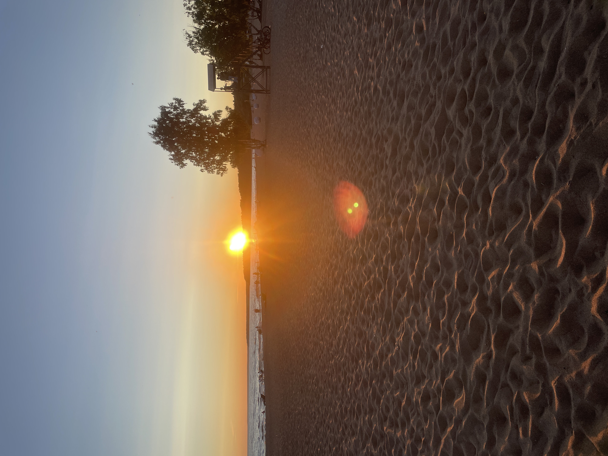 A wide-angle, eye-level shot captures a sandy beach during sunset in Solnechnoye, Russia. The sun, a bright orange orb, sits low on the horizon, casting a warm glow across the scene. Its rays create a lens flare in the foreground, with two distinct green orbs visible. The foreground is dominated by textured sand, showing ripples and patterns from the tide or wind. In the midground, the gentle waves of the Gulf of Finland meet the shore. Several figures are scattered along the shoreline, some appearing to walk or stand near the water's edge, while others are seated on chairs or lying on lounge chairs. Further inland, a tall lifeguard tower with a roof structure stands prominently, with at least two individuals visible on its platform. Two bicycles are parked near the tower. Trees, including a notably large one to the left of the tower and others on the right, flank the beach. The sky transitions from a pale yellow at the horizon to a clear blue overhead, with a few faint, small dark specks, possibly birds or insects, visible against the sky. The overall atmosphere is serene and depicts a typical summer evening at a Russian coastal town.
