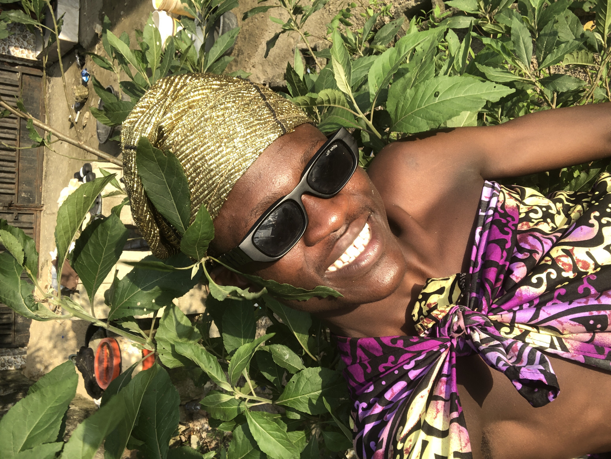 A person is lying down amongst dense green foliage. The person is wearing a gold-colored headwrap and black sunglasses. They have a bright smile and their teeth are visible. Around their neck and shoulders, they are wearing a brightly colored, patterned fabric tied in a knot. The background appears to be a mixture of dirt, gravel, and some debris, possibly an outdoor area in Ado, Nigeria. A metal fence can be seen in the upper background.