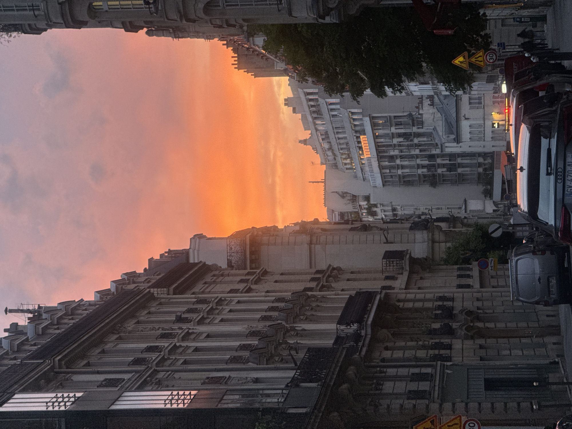 Here's a description of the image:
The image captures a street scene in Paris, France, at sunset. The sky is filled with hues of orange and pink, illuminating the architecture. Tall buildings with ornate facades line the street, characteristic of Parisian architecture. Several cars are parked along the side of the street. Traffic signs are also visible. Trees are visible along the right side of the buildings. The image is rotated to the left.