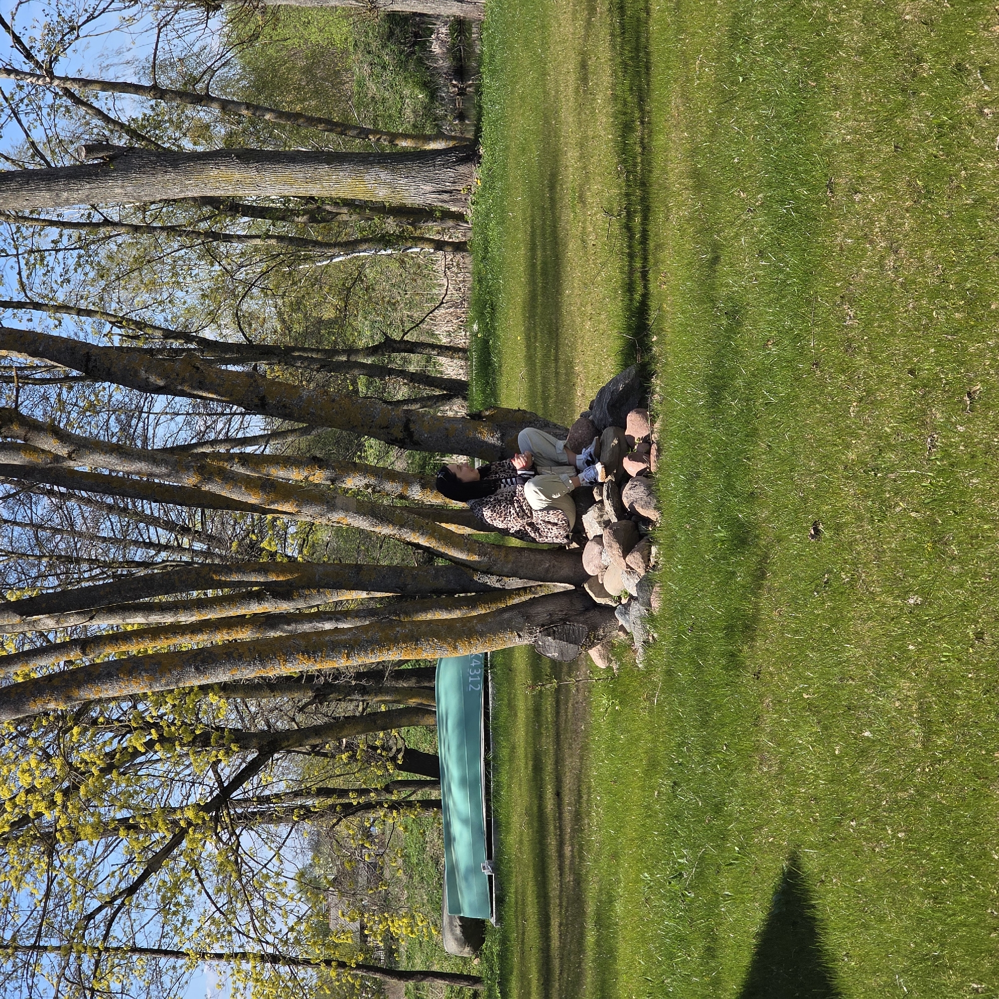 A woman is lying on a stack of rocks near a tree in Капцёўскі сельскі Савет, Belarus. The woman has dark hair and is wearing a leopard print jacket, a striped shirt, and light-colored pants. A canoe is leaning against the tree to the left of the woman. The ground is covered in green grass and there are trees in the background.