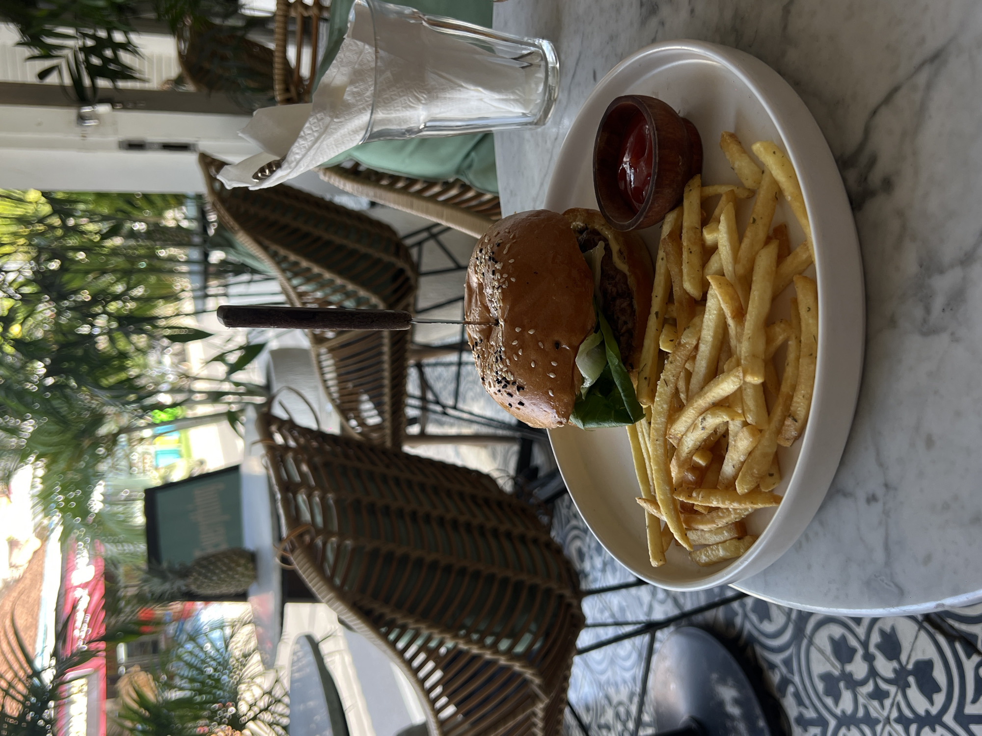 The image shows a burger and fries meal served on a white plate, accompanied by a small wooden bowl of ketchup. A knife is piercing the burger. A glass filled with napkins is next to the plate on a marble-patterned table. The scene appears to be in an outdoor dining area with woven chairs and greenery visible in the background. The location is identified as North Kuta, Indonesia. There are no people visible in the image.