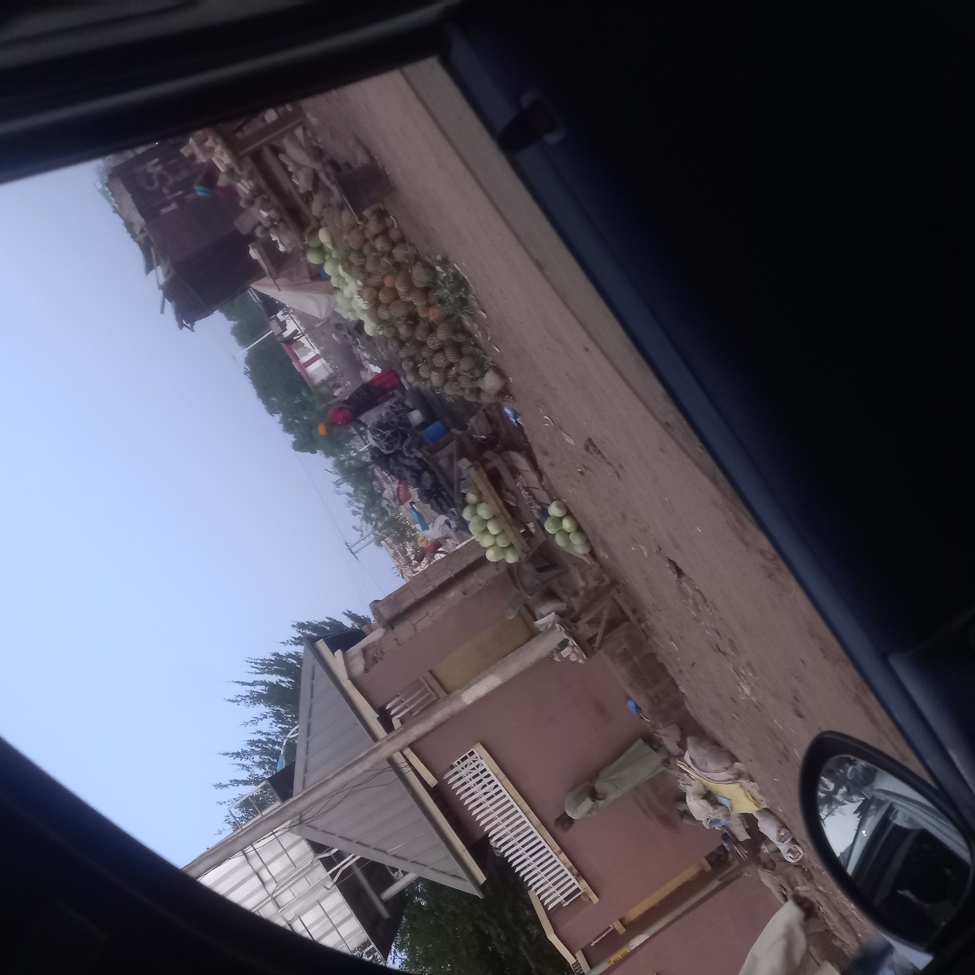 View from a car window onto a street scene in Kinkinau, Nigeria. The street displays piles of watermelons and pineapples for sale. A person is visible near a building with a partial metal roof. The landscape consists of dusty roads and buildings. The presence of vegetation and power lines indicate an urban or suburban setting.