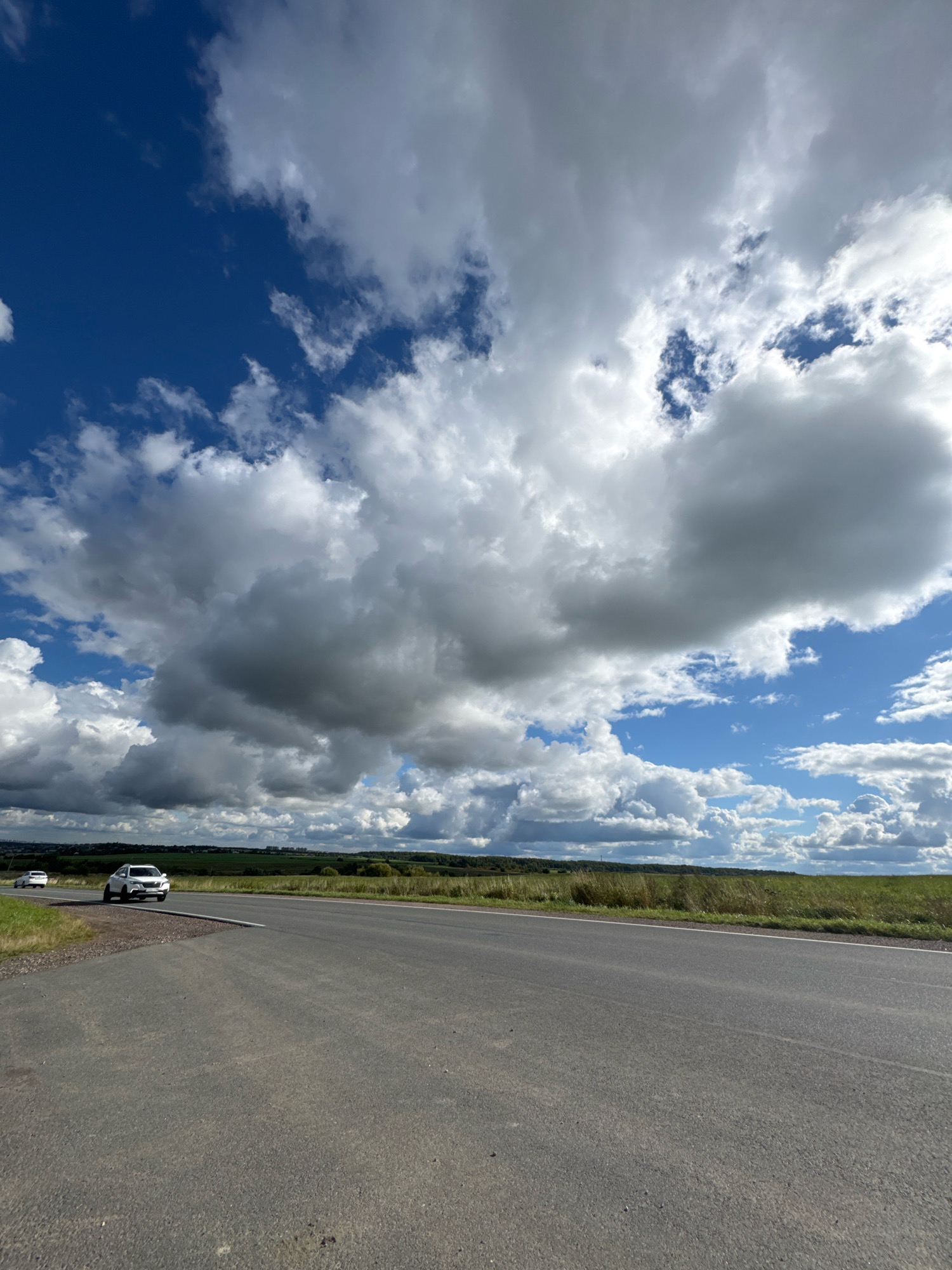 The image captures a rural scene in Kaporskoe, Russia, featuring a two-lane asphalt road curving to the left. Two white cars are visible on the road, one in the foreground and another further in the distance. The landscape is characterized by grassy fields and distant trees under a dramatic sky filled with large, white, and gray cumulus clouds against a bright blue background.