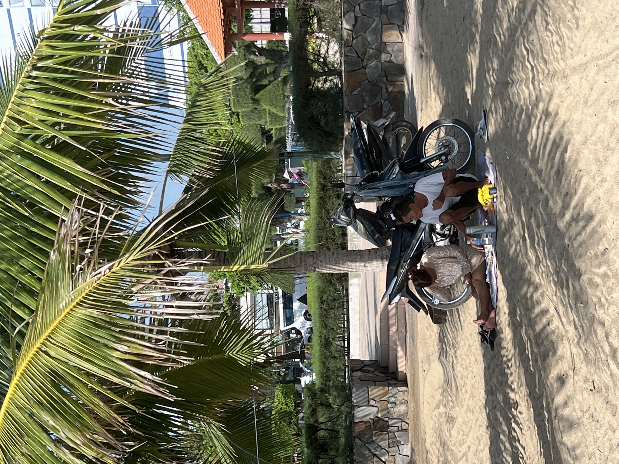 This image depicts two individuals sitting on a sandy beach in Nha Trang Ward, Vietnam, beneath the shade of a palm tree. In the background, there are two motorcycles parked, one of which has a denim jacket draped over it. Beyond the motorcycles, a stone wall with a paved walkway and steps is visible, leading to a landscaped area with manicured bushes and trees. Further in the background, a red-roofed pavilion and a tall, modern building can be seen. The individuals appear to be having a meal or snack, with food items and drinks on a mat. One person is looking at their phone, while the other is facing away from the camera. The sand is casting shadows from the palm fronds.