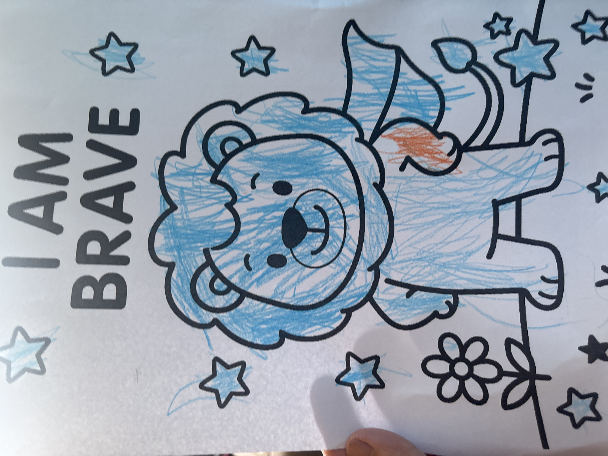 This is a photograph of a coloring page featuring a cartoon lion with the words "I AM BRAVE" written above it. The lion is colored in blue with orange accents on its cape. There are stars and a flower drawn around the lion. The coloring page is on a white surface, and a person's hand is visible on the left side of the image, holding the page. The location of this image is not discernible from the provided visual information; therefore, it cannot be definitively placed in Dubai, United Arab Emirates.