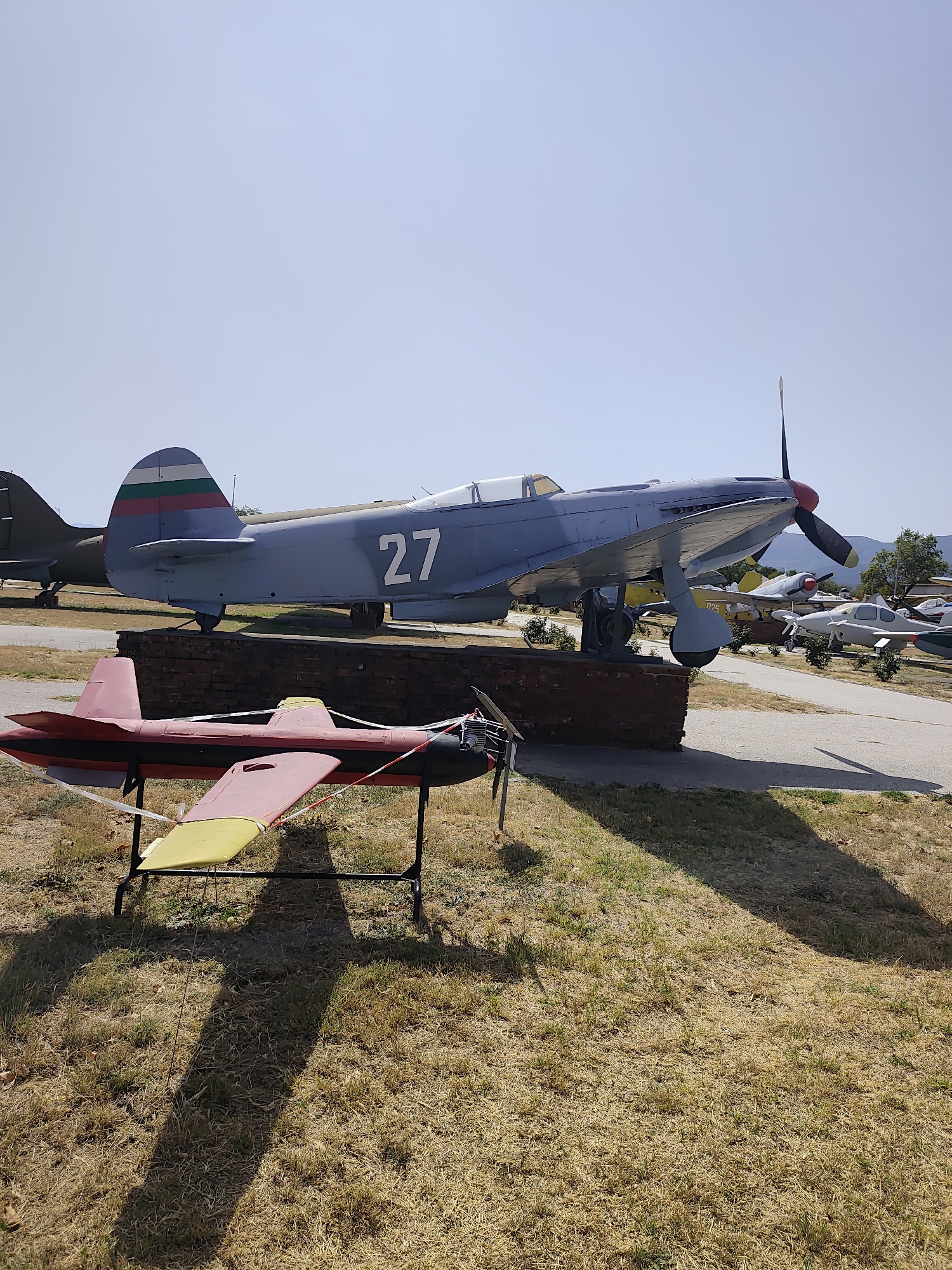 In the city of Krumovo, Bulgaria, a scene unfolds in an outdoor aviation museum or display area. In the foreground, a small, red and yellow model airplane with a propeller is mounted on a black stand, casting a long shadow across the dry, grassy ground. Behind it, a larger, light blue military aircraft with the number "27" prominently displayed on its side rests on a brick pedestal. The tail fin of this aircraft features the Bulgarian flag. The background reveals several other aircraft, also on display, with a clear, bright sky overhead and distant hills or mountains. The overall environment appears to be an open, sunny area, possibly a disused airfield or a dedicated museum ground.