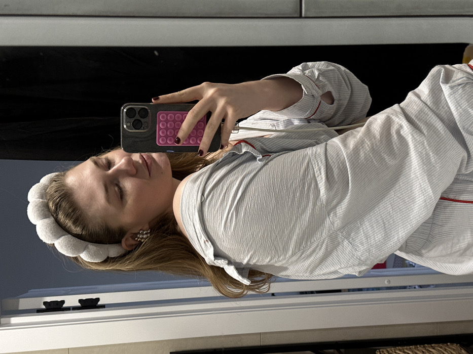 The scene is located indoors in the city of Bakcha-Sarai, Russia. An adult female is depicted from the upper torso upwards, taking a mirror selfie with a smartphone held in her right hand. She has light brown hair styled down and wears a light grey, segmented headband. Her left ear displays a silver-toned, multi-clustered earring. She is dressed in a light-colored, striped pajama-style top with red piping, which is pulled off her left shoulder. Her eyes are closed or looking downwards, and her head is slightly tilted. The smartphone is dark-colored, featuring a triple-lens camera array and a pink pop-it style accessory adhered to its back. A white cable extends from the phone. The background visible in the mirror includes a white door frame with dark hinges on the left, a dark surface behind the subject, and a light grey tiled or paneled wall section on the right. A faint reflection of a woven basket is discernible in the lower-left corner near the door frame.