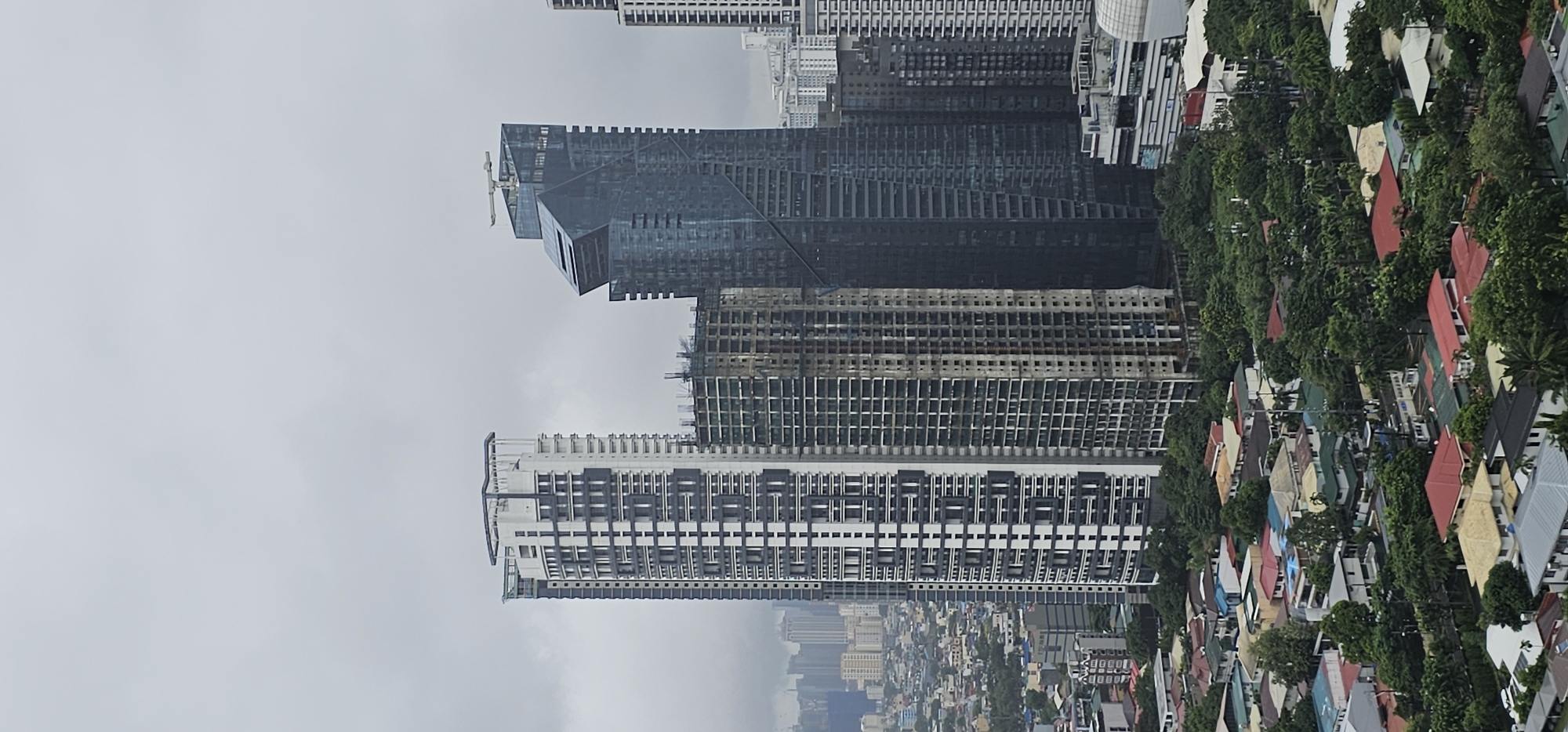 This is a vertical, eye-level shot of a cityscape in Makati, Philippines, under an overcast sky. The scene is dominated by several tall buildings, some under construction and others completed.
In the foreground, a cluster of residential houses with red and other colored roofs are visible, interspersed with lush green trees. Behind these houses, the construction of a medium-rise building is apparent, with its skeletal framework exposed.
Further back, three prominent skyscrapers stand out. The leftmost is a tall, modern white and gray building with a repetitive pattern of windows and balconies. To its right is another skyscraper that appears to be under construction, showing exposed concrete and rebar. The third skyscraper, located in the center-right of the image, is characterized by its dark, reflective glass facade and an unusual, angular design. A crane-like structure is visible at its peak.
In the distance, a denser collection of buildings suggests the heart of the city, with more skyscrapers and structures fading into the hazy sky. The overall impression is one of urban development and ongoing construction in a tropical environment.
