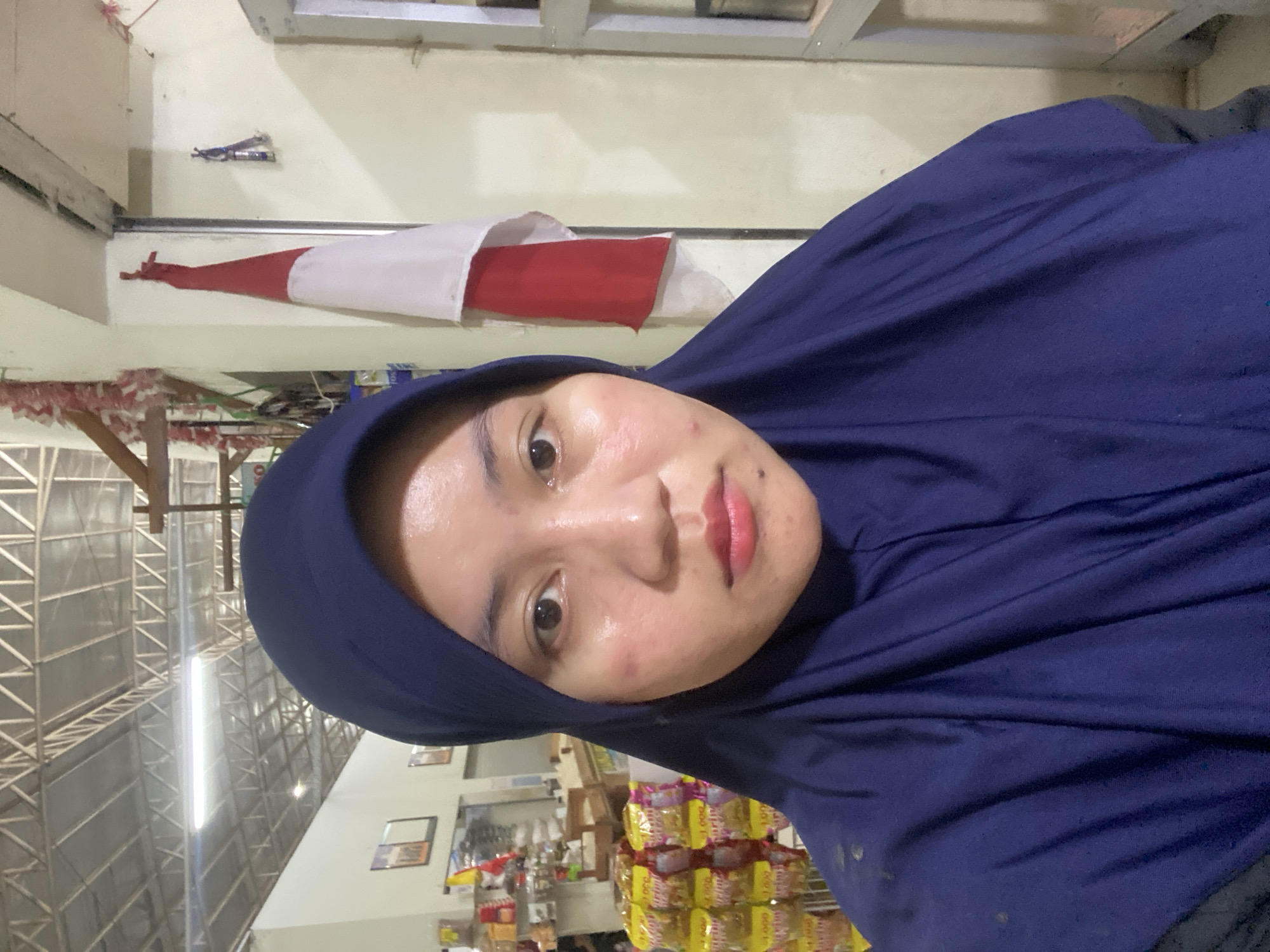 A selfie of a young woman wearing a dark blue hijab is taken indoors, likely in a shop or market, judging by the shelves of products visible in the background. The woman is looking directly at the camera. She has fair skin with some redness on her cheeks and a few blemishes. Her lips are painted a light pink. Behind her, on the wall, hangs a red and white Indonesian flag. The ceiling above appears to be industrial with exposed metal beams and lighting. The overall lighting is bright, suggesting artificial illumination.