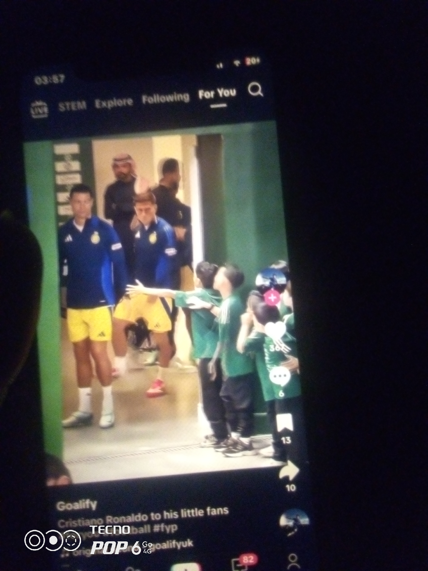 The image shows a mobile phone screen displaying a video. The video features Cristiano Ronaldo and other individuals, some appearing to be young fans in green shirts. The setting seems to be a stadium tunnel or hallway. On the phone screen, a timestamp "03:57" is visible at the top left, followed by navigation options "LIVE", "STEM", "Explore", "Following", and "For You". A search icon is also present. Below the video, text reads "Goalify Cristiano Ronaldo to his little fans Tecnoball #fyp" and "Go@goalifyuk". A logo with "POP 6" and "TECNO 4G" is also visible at the bottom. The overall lighting suggests the video is being watched in a dimly lit environment. There is no information in the image indicating the location as Howayi, Nigeria.