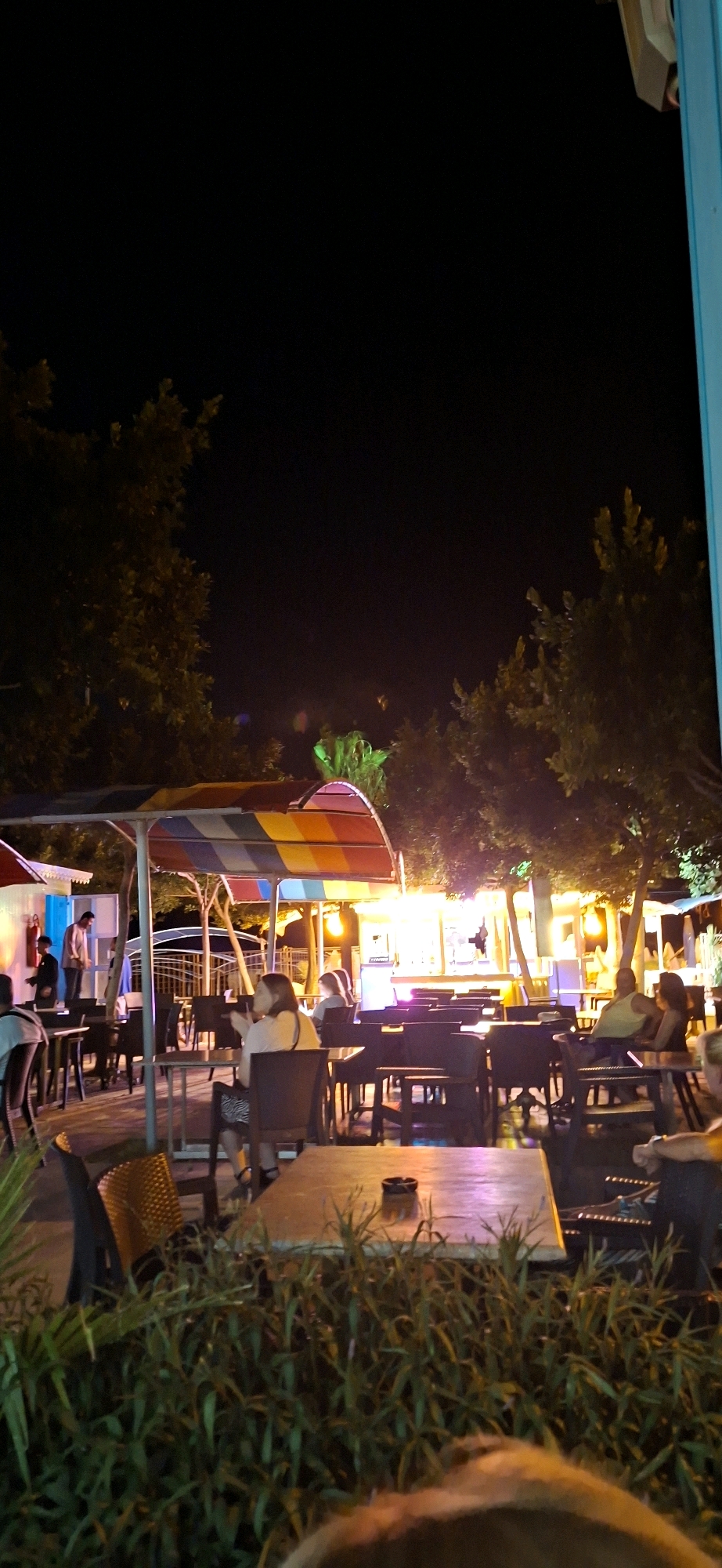 This image captures an outdoor seating area of a restaurant or cafe at night, likely in Kemer, Turkey. A brightly colored canopy, featuring stripes of red, orange, yellow, and blue, is visible over some of the tables and chairs. The scene is illuminated by artificial lighting, casting a warm glow on the restaurant's facade in the background, which appears to have large windows or open spaces. Several tables and chairs are arranged throughout the outdoor space, with some occupied by patrons. In the middle ground, a woman with dark hair is seated at a table, looking to her left and holding a drink. To her right, another woman is also seated at a table, her face partially obscured. In the background, near the building, two individuals are standing. One, a man in a light-colored jacket, is looking towards the other person. Trees with dense foliage are present throughout the scene, with some branches extending over the seating area. The ground appears to be paved. In the foreground, a patch of green vegetation, possibly ornamental plants or bushes, partially obscures the view of the tables and chairs closest to the camera.