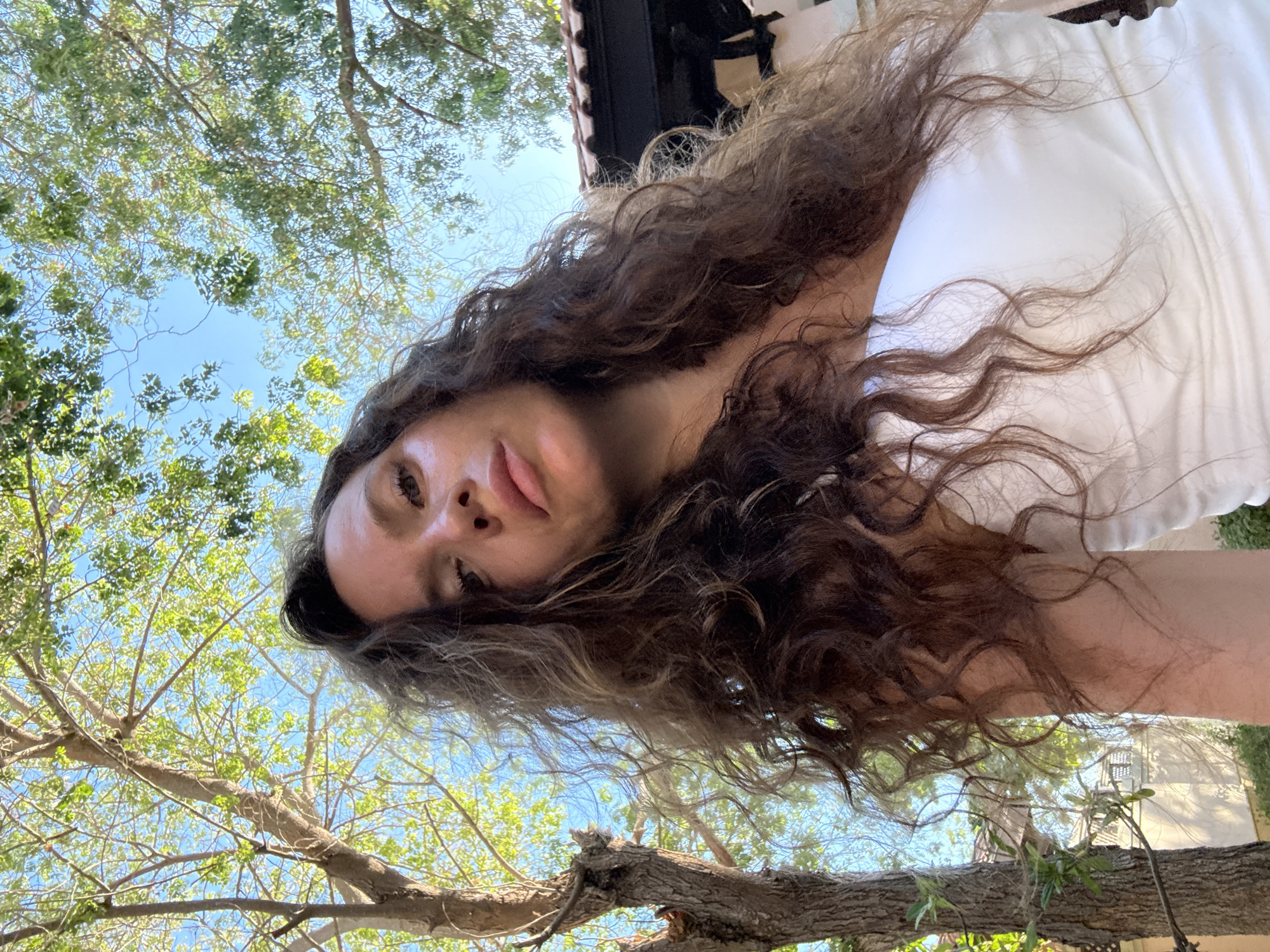 A woman with long, curly brown hair and fair skin is shown from the chest up, looking at the camera with a serious expression. She is wearing a white strapless top. The background is filled with green trees and a bright blue sky, suggesting an outdoor setting. A section of a tiled roof and a building are visible in the upper right portion of the image, and a tree trunk is to the left. The overall lighting indicates it is daytime, possibly sunny given the highlights on the woman's skin. The scene is likely a garden or park area. There are no discernible features that place this image specifically in Dubai, United Arab Emirates.
