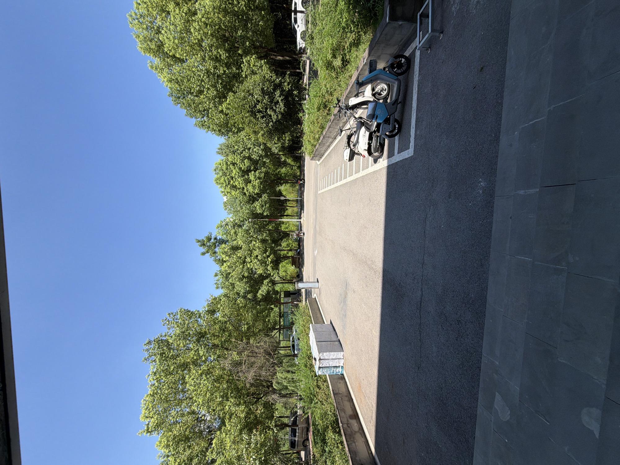 The image depicts an outdoor paved area, likely a parking lot, under a clear blue sky. Lush green trees line the background, with some foliage bordering the paved area. In the foreground, a dark gray tiled surface is visible. On the paved area, a stack of white, shrink-wrapped rectangular objects, possibly building materials, sits near a low concrete wall. Further back, two electric scooters, one white and one blue, are parked in designated white lines. Several vehicles, including a silver minivan and a white car, are parked in the distance, as are streetlights and some signage. The scene appears to be a quiet, daytime setting in a public outdoor space, potentially in Jiangjin District, Chongqing, China, given the presence of the scooters and the overall landscape.