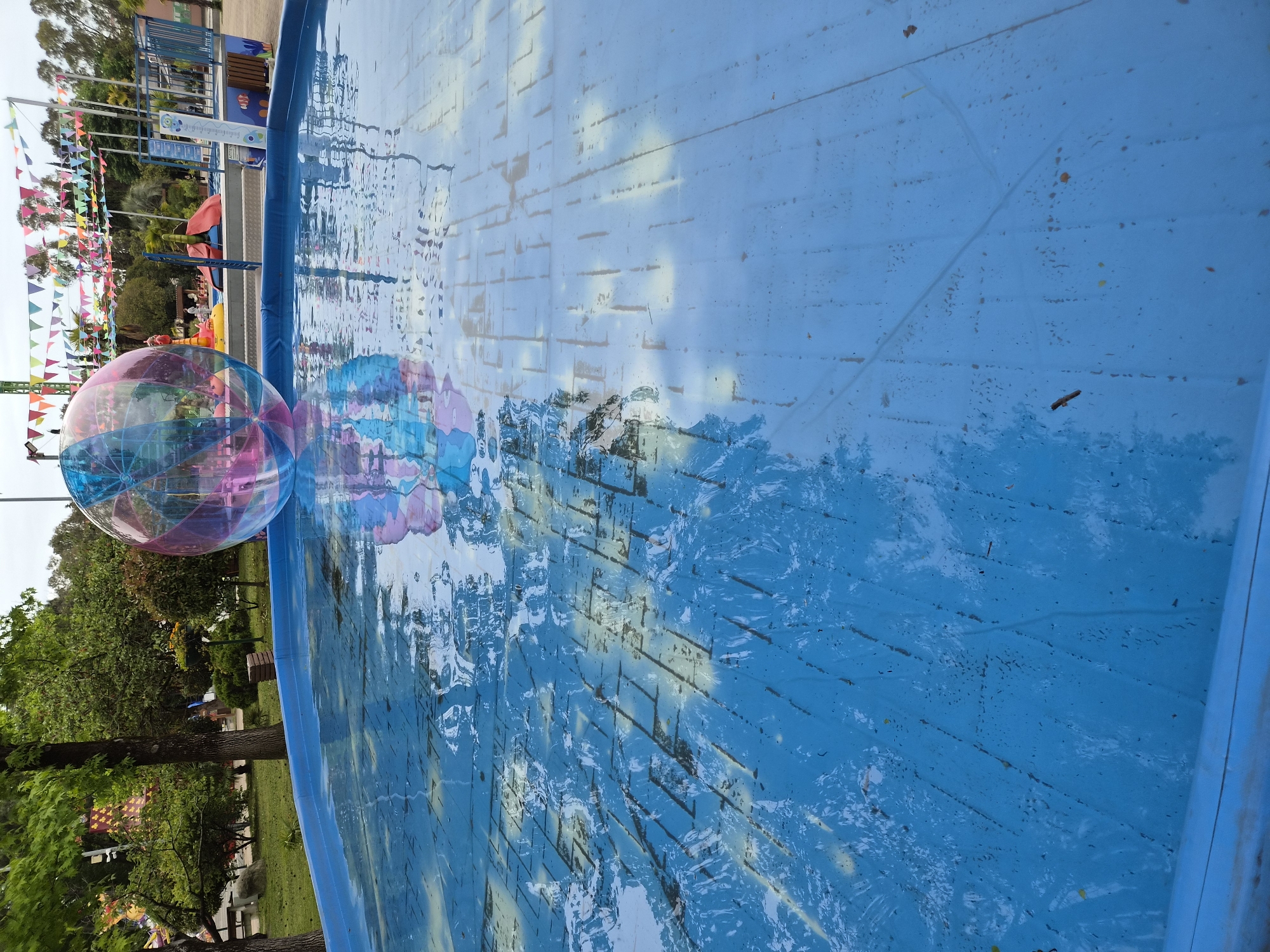 The image depicts a water-filled, blue-painted pool or fountain at an outdoor venue. Two large, clear inflatable balls with colorful accents are resting on the edge of the pool, partially submerged in the water. The surface of the water reflects the surrounding environment, including trees, flags, and the sky. A blue railing borders the pool. Trees are visible on the left side, and colorful flags are strung up in the background, along with some park facilities. Based on visual cues in the broader scene, this image is located in Sochi, Russia.