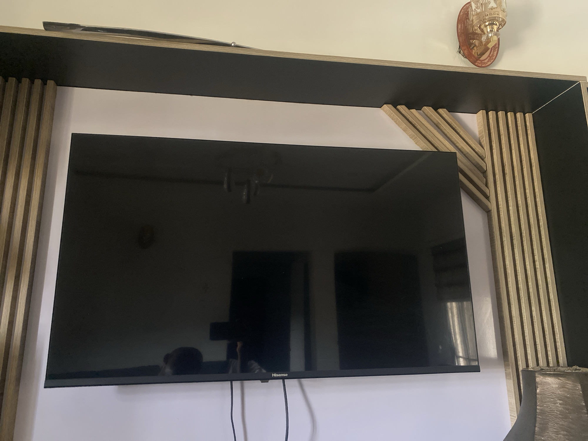 This image captures an indoor living space in Abuja, Nigeria, featuring a prominent media setup. The main subject is a large, black, flat-screen television, visibly branded "Hisense" at the bottom center. The TV screen is off and highly reflective, mirroring elements of the room, including a ceiling light fixture and dark rectangular shapes that appear to be doorways or windows.
The television is integrated into a custom-built media console or entertainment unit. This unit is characterized by dark gray or black horizontal panels forming shelves or framing, contrasted with light wood-toned vertical slats. On the right side of the TV, some of these wooden slats are arranged at an angle, creating a dynamic design element. The wall directly behind the TV and within the console's structure appears to be a light color, possibly white or very light gray.
Above the media unit on the right side, a decorative wall sconce light fixture with a gold or bronze base and a textured glass shade is mounted. In the bottom right corner of the frame, part of a lamp or decorative item with a dark, textured lampshade is visible. Wires are seen hanging vertically below the television. The overall decor suggests a modern and styled interior. No people are directly visible, though reflections in the TV hint at the presence of the photographer and other elements in the room.
