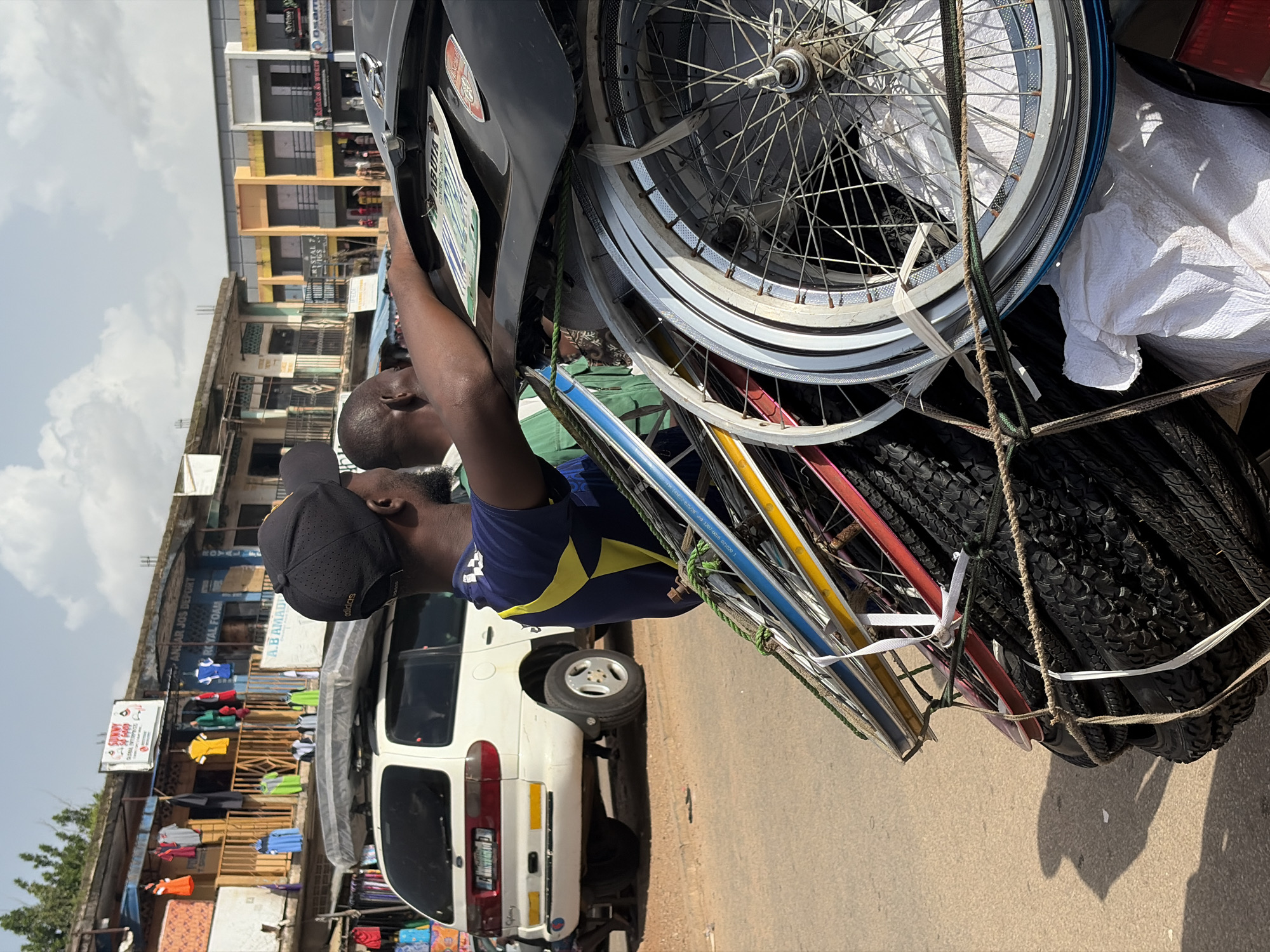 In the city of Jos, Nigeria, two men stand near a white Ford Galaxy minivan. The minivan is loaded with various items, including a mattress on its roof and a significant stack of bicycle wheels and tires secured to its rear. One man, wearing a black baseball cap and a dark blue shirt with yellow accents, has his arm resting on the vehicle's hood, near a license plate with "ABUJA" printed on it. The other man, in a green shirt, stands partially behind him. In the background, a row of shops with clothing displays and signage, including one that reads "SUNNY GOLD GLOBAL ENTERPRISES," lines the street. The sky is partly cloudy.