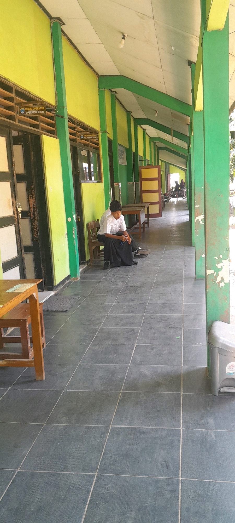 A young male student wearing a white t-shirt and a black sarong sits on a wooden chair in an outdoor corridor of a school in Tahunan, Indonesia. He is looking down at something in his hands. The corridor has a yellow wall on the left and green pillars supporting a green roof. The floor is tiled with dark gray squares. Two signs are visible on the yellow wall: one reads "RUANG OPERATOR OPERATOR ROOM" and the other reads "RUANG KEPALA SEKOLAH PRINCIPAL ROOM". Further down the corridor, a wooden table and chairs are visible, and in the background, two people on motorcycles are seen exiting the school premises. To the right of the corridor, a gray trash can is visible. The scene appears to be a quiet moment at a school, possibly during a break or after school hours.