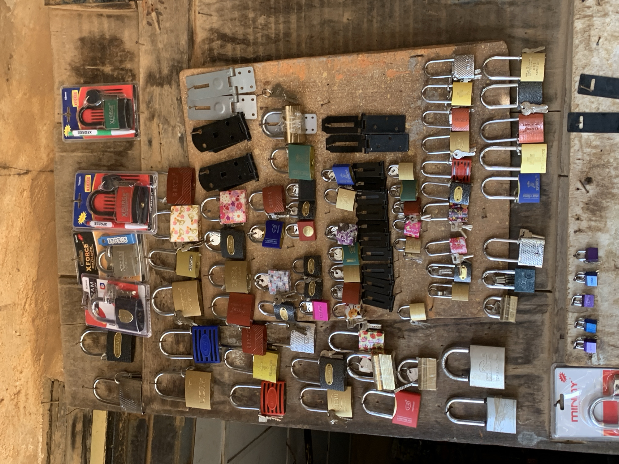 A collection of padlocks of varying sizes, colors, and brands (XForce, Zam, Liand, Renda, Miny) are arranged on a weathered wooden surface. Some padlocks are new in their packaging while others are loose, with keys attached to some. The scene could be a vendor's display or a storage area, possibly in a market setting, due to the assortment and arrangement of the locks. Based on the presence of a "MINY" brand padlock package in the bottom right corner and the presence of padlock packages with "XFORCE" branding on the left side, the scene may be a hardware or lock shop located in Ikorodu, Nigeria. There are no people visible in the image.