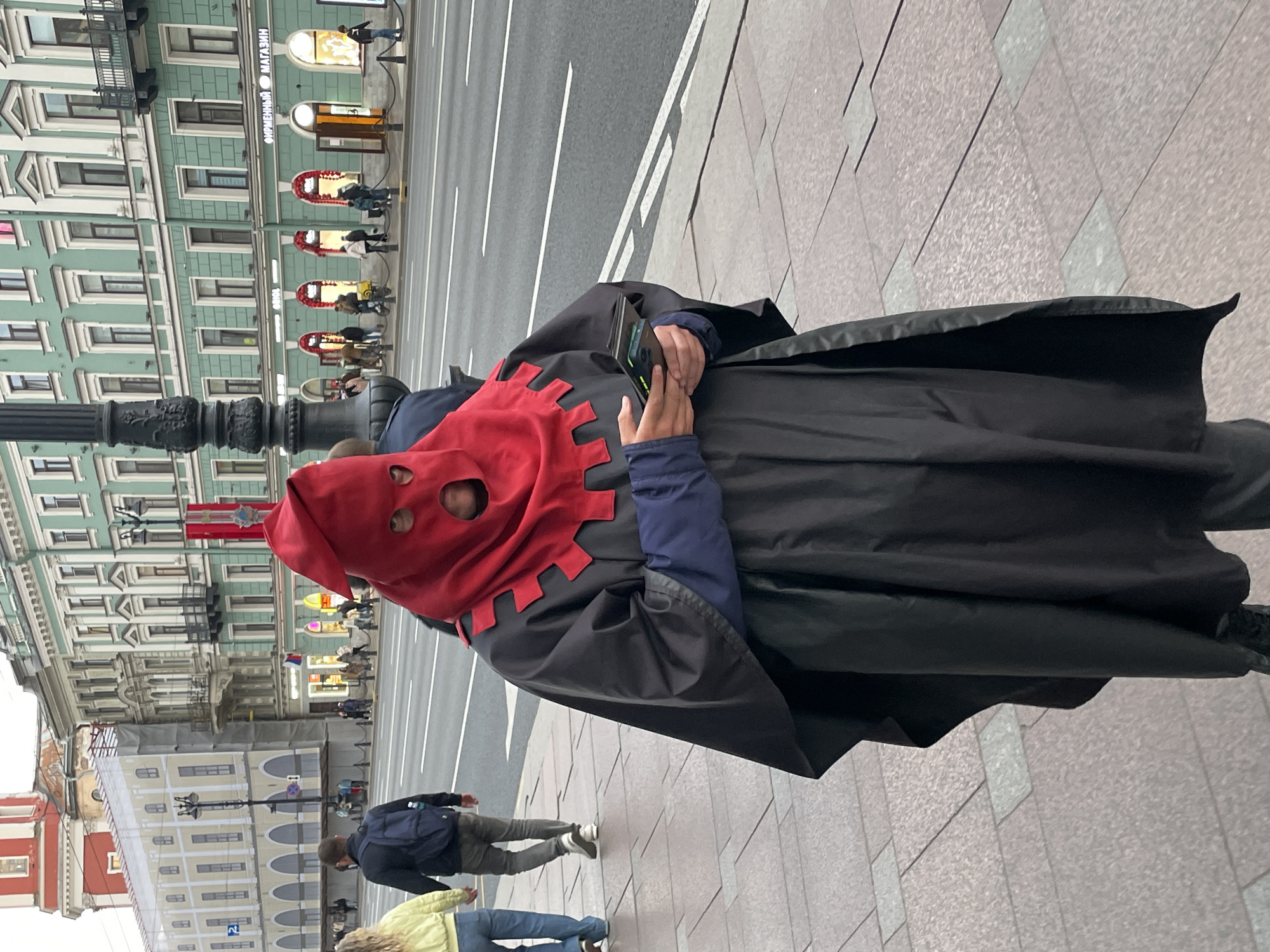 A person wearing a red hood with eye holes and a black cloak stands on a sidewalk, holding a smartphone. The person is facing right and is slightly off-center to the right.
In the background, across a street, there are several buildings. A tall, green building with white trim and multiple windows is visible, along with other buildings with architectural details. A lamppost stands to the left of the person, and other pedestrians are walking on the sidewalk and the crosswalk in the background.
The location appears to be in Saint Petersburg, Russia, based on the architectural style and the Cyrillic text on a storefront.