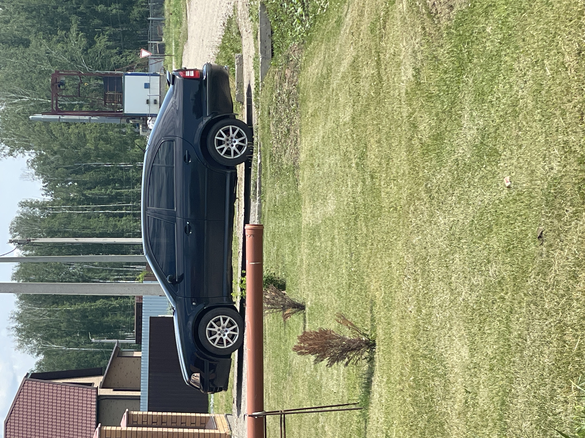 A black Skoda Octavia is parked on a gravel and grass area in Chulym, Russia. The car is facing left and has tinted windows. In the background, there is a brick house with a red tiled roof, a dark fence, and a tall industrial structure with a red frame. Trees form a dense line behind the industrial structure. A reddish-brown pipe is laid horizontally across the foreground, supported by a metal frame. Several small, dry-looking bushes are scattered on the green grass in front of the pipe. The overall environment appears to be a rural or semi-industrial area on a bright, sunny day.