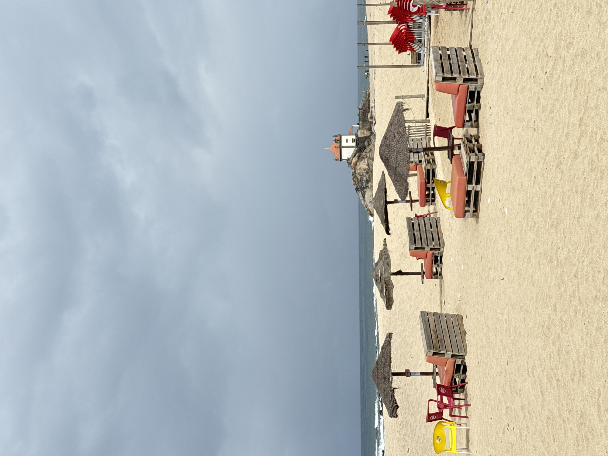 The scene is a beach in Vila Nova de Gaia, Portugal, on a cloudy day. The view is of the shoreline from an elevated position, looking towards a white and orange chapel sitting atop a rocky cliff. Scattered along the beach are several thatched beach umbrellas and beach furniture constructed from wooden pallets and colorful plastic chairs. A stack of folded red beach umbrellas sits farther up the beach. The sand is light-colored, and the sea is visible along the coastline. The sky is overcast. There are no people immediately visible in the image.