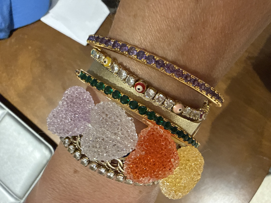 A person's wrist is adorned with a stack of bracelets. The top bracelet is gold with purple stones. Below that, a gold bracelet features clear stones and eye-shaped charms. The third bracelet is gold with green stones. At the base, a wider gold bracelet supports four translucent, beaded hearts in lavender, white, orange, and yellow. The bottom bracelet has a gold base with clear stones.