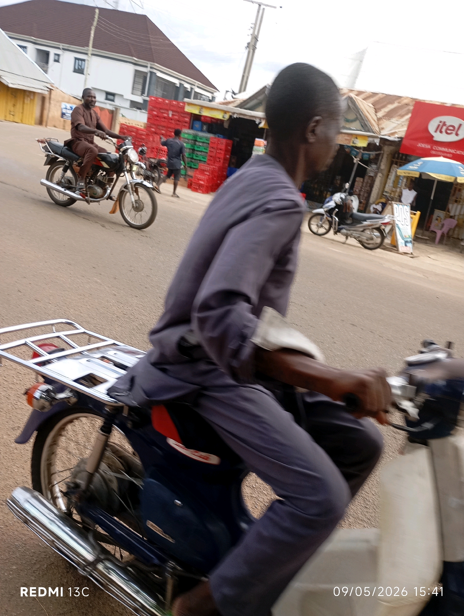 In Shendam, Nigeria, a man rides a motorcycle on a paved road. He is wearing a dark blue tunic and trousers. Another man on a similar motorcycle is visible further down the road. In the background, there are buildings, including a red-roofed structure with a sign that reads "itel". Stacks of red and green crates are also visible. The scene appears to be a busy market or commercial area.