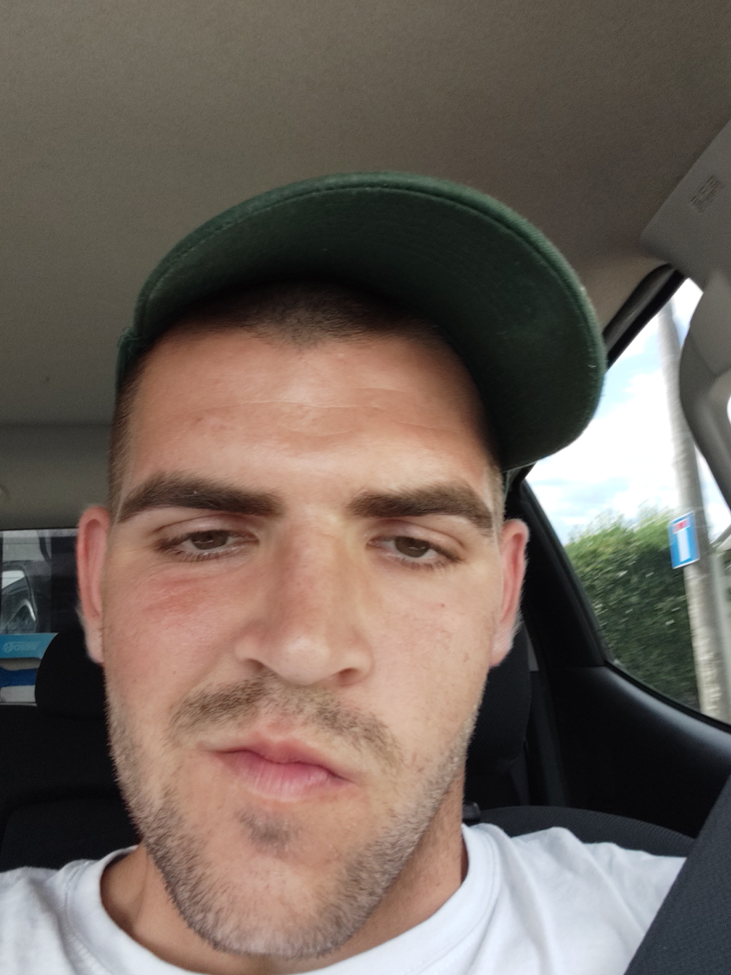 The image is a selfie of a man inside a car, likely in Lombardore, Italy. The man is wearing a green baseball cap and a white t-shirt. He has short, dark hair, brown eyes, and a light stubble on his face. His lips are slightly parted. The background shows the interior of a car, with a glimpse of the outdoors through the window, suggesting it's daytime. Trees and a road sign are visible outside, indicating a rural or suburban setting. The man's expression is neutral. There is no visible text in the image.