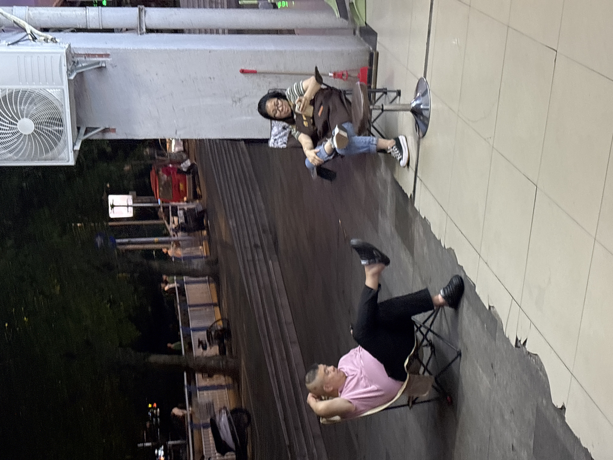 The image captures an outdoor scene at what appears to be night or dusk, showing two individuals relaxing. The photograph itself is rotated, making the subjects appear to be on a vertical surface.
In the left half of the frame, a woman is seated on a tall, chrome-based swivel stool. She wears glasses, a striped t-shirt under a brown apron, ripped blue jeans, and black and white sneakers. She holds a black mobile phone in her lap, smiles, and looks directly towards the viewer. Behind her is a concrete wall with an air conditioning unit mounted on it, and several pipes running horizontally. A red pole leans against the wall nearby.
To the woman's right, a man is reclined on a black portable folding chair. He wears a pink t-shirt and black pants with black sneakers. His hands are clasped behind his head, and one leg is propped up. He appears to be resting comfortably.
The ground surface transitions from light-colored tiles beneath the woman to a darker, rougher, and partially broken concrete or asphalt area where the man is situated. In the background, beyond a set of steps or terraced seating, dim lights, trees, and dark structures with railings are visible, along with a faint red object that might be a vehicle. No discernible text is visible anywhere in the image. The overall impression is one of casual relaxation in an urban or semi-urban outdoor setting in Nan'an District, China.