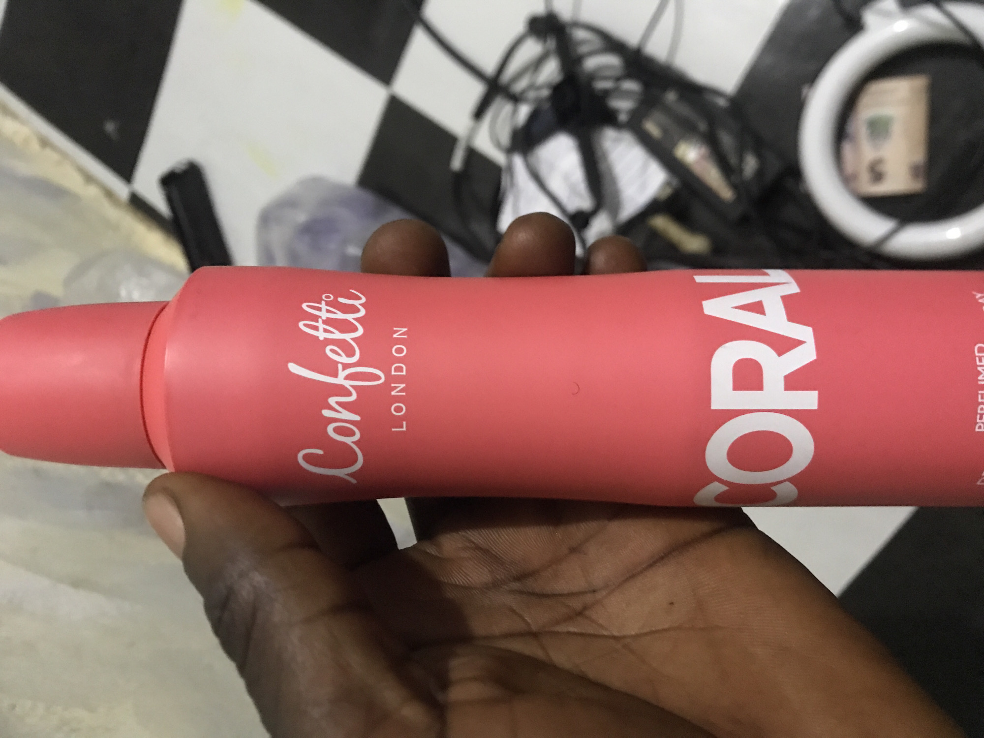 This forensic description details an indoor scene in Rafin Par, Nigeria. A dark-skinned human hand, positioned on the left, firmly grasps a coral-pink aerosol can, holding it vertically. The can features "Confetti" in white script font near the top, followed by "LONDON" in smaller white capital letters. Below, the word "CORAL" is prominently displayed in large, bold white capital letters. Partially visible text beneath "CORAL" reads "PERFUMED" and then "SAY". The background on the right reveals a black and white checkered floor. Scattered on the floor are various electronic cables and a black object resembling a power strip or adapter. In the lower right corner, a white ring light is visible, with what appears to be a Nigerian Naira banknote (possibly a 500 Naira note) resting within its circular frame. The background on the upper left consists of a light-colored, textured surface.