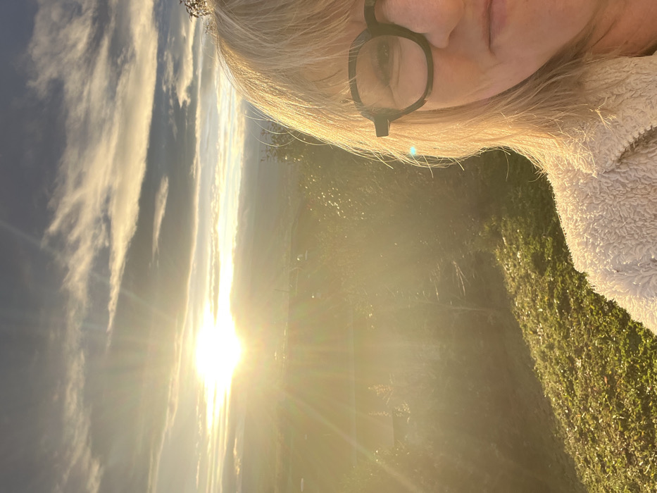 A selfie during sunset, featuring a person with blonde hair and glasses. The sun is low on the horizon, creating a bright glare and lens flare. Clouds are visible in the sky. The person wears a fuzzy jacket and the background includes some grass and vegetation, adding a natural element to the scene. The lighting is very warm due to the sunset.