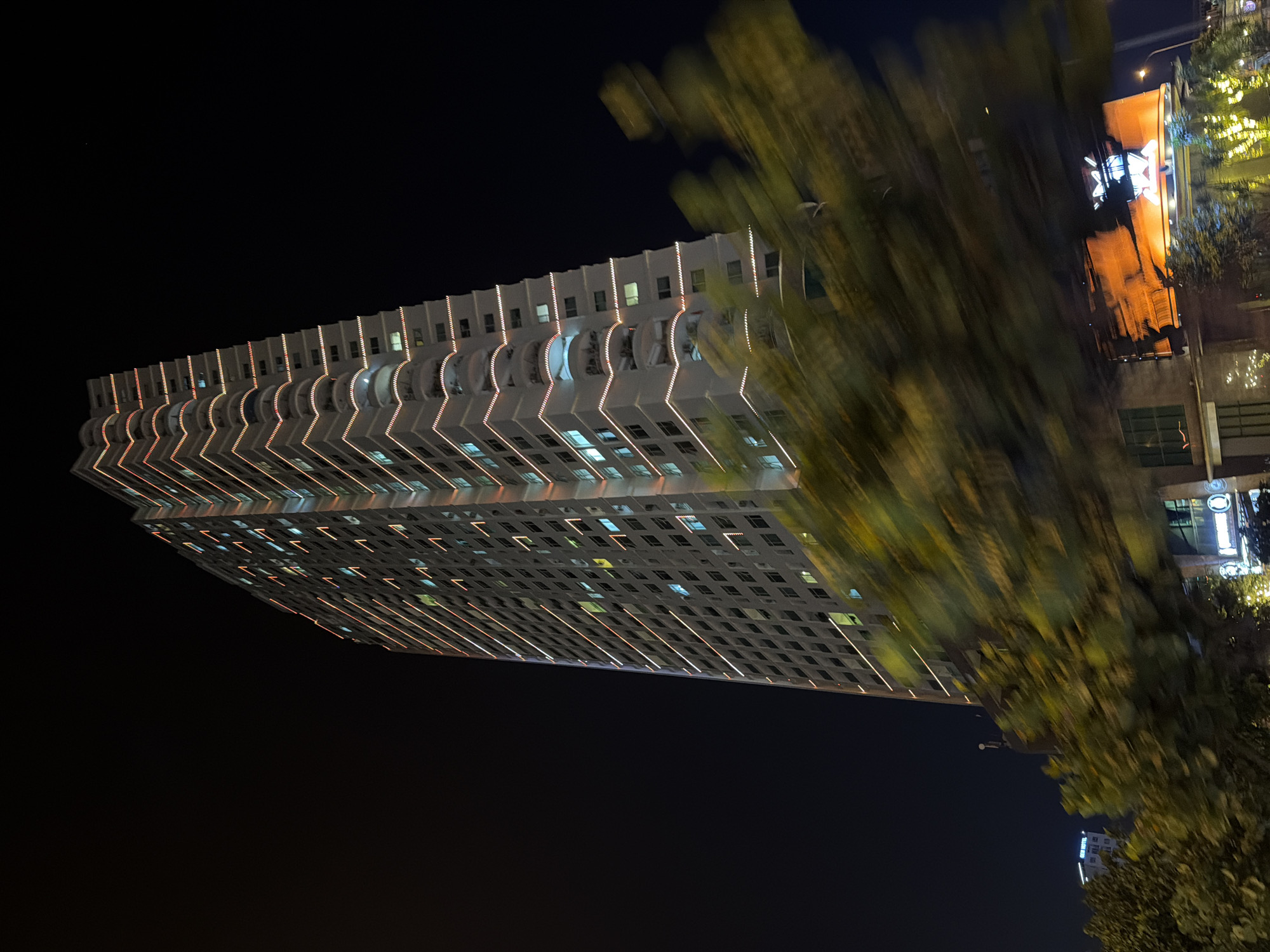 The image displays a nighttime low-angle view of a prominent, multi-story building, likely a residential or hotel high-rise, situated in the city of Bac Nha Trang Ward, Vietnam. The building's light-colored facade is illuminated by numerous interior lights visible through its windows, some appearing blue-white and others yellow-orange. Decorative warm-toned LED strip lighting outlines horizontal elements on the left side of the building and accentuates the distinct wavy or undulating balconies on the right side. The tall structure rises into a dark, starless night sky.
The immediate foreground features a dense mass of green and yellow foliage, heavily blurred horizontally, indicating the photograph was taken from a moving perspective. Below this blurred vegetation, parts of lower buildings and various street-level lights, including a large, indistinct orange-lit sign on the bottom right, are visible. In the distant left background, a smaller building with a blue illuminated sign reading "GEN HOTEL" can be observed. No individuals are clearly identifiable, and no specific actions or interactions are depicted within the frame.