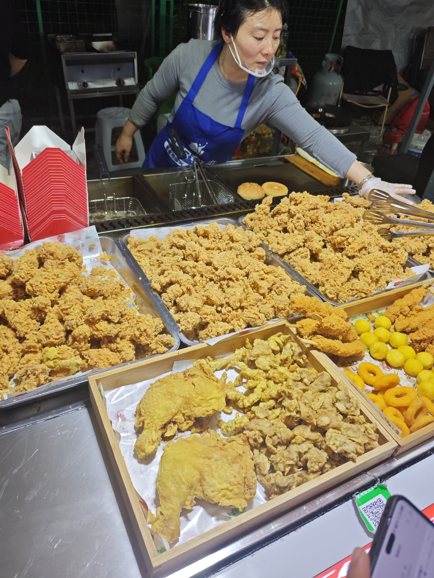 The image shows a street food vendor in Shapingba District, China. A woman wearing a grey long-sleeved shirt, a blue apron, and a transparent face shield is tending to a display of various fried foods. The foods include fried chicken pieces, donut-shaped fried items, skewers of fried meat, and small fried potato balls. Metal trays and wooden boxes hold the food. Behind the vendor, there is cooking equipment, including fryers and a griddle with buns on it. Red boxes are stacked on the left. In the lower right corner, a hand holds a smartphone that is pointed towards a green QR code.