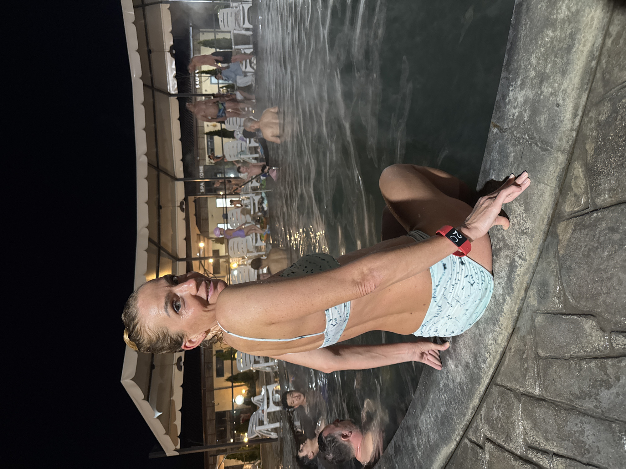 An outdoor night scene depicts people relaxing in a thermal pool at Kara-Oy, Kyrgyzstan. In the foreground, a woman with wet blonde hair, wearing a light blue patterned bikini, sits on the dark, stone edge of the pool. She smiles and looks towards the camera, her left wrist adorned with a red smartwatch displaying "20." Steam rises from the dark water, indicating its warmth.
Several other individuals are visible in the background, mostly submerged in the pool or seated on its perimeter and nearby white plastic chairs. The general ambiance is one of leisure and social gathering. Behind the pool, illuminated structures with white awnings and string lights suggest a resort or spa facility. Blurry signs in the background show text such as "SAUNA" and "SPA ZONE," confirming the setting as a spa or thermal complex. The sky is dark, establishing the nighttime environment.