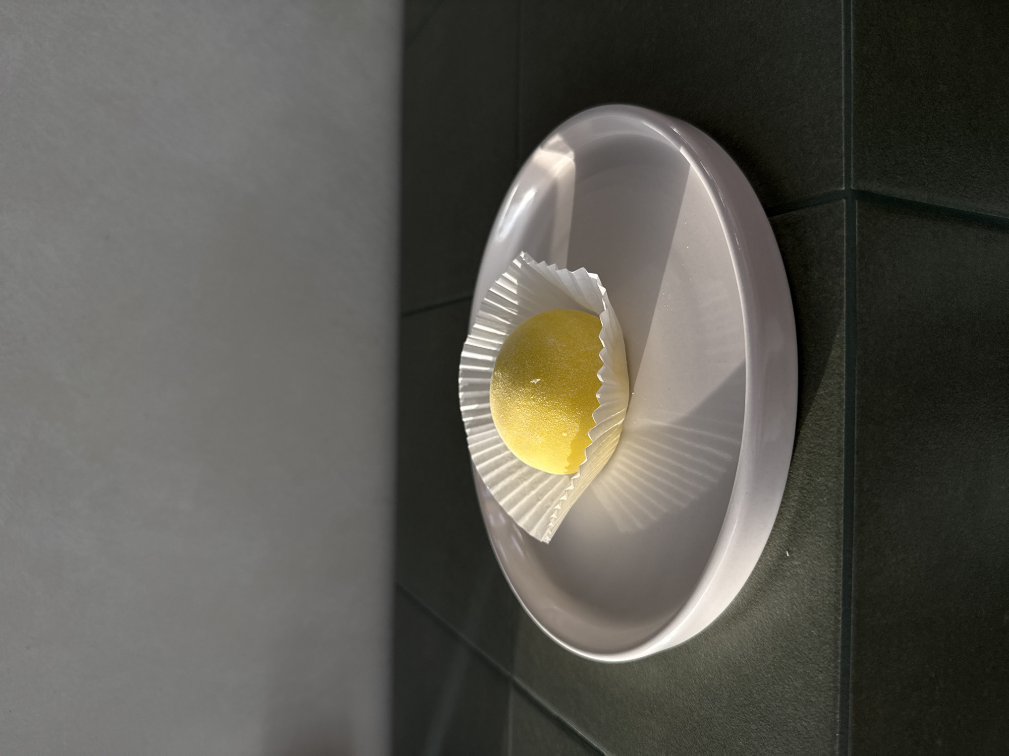 A single, bright yellow, spherical confection, possibly a mochi or cake, rests on a white, circular plate. The confection is nestled within a fluted white paper liner. The plate is positioned on a surface with dark, textured tiles that appear to be part of a wall or countertop. To the left of the plate, there is a smooth, gray surface that reflects a diagonal band of light, suggesting the presence of a window or strong light source. The lighting creates distinct shadows on the plate, indicating it is daytime, likely morning or afternoon, with sunlight streaming in. The setting appears to be indoors, possibly a kitchen or a minimalist cafe. There is no visible text in the image.