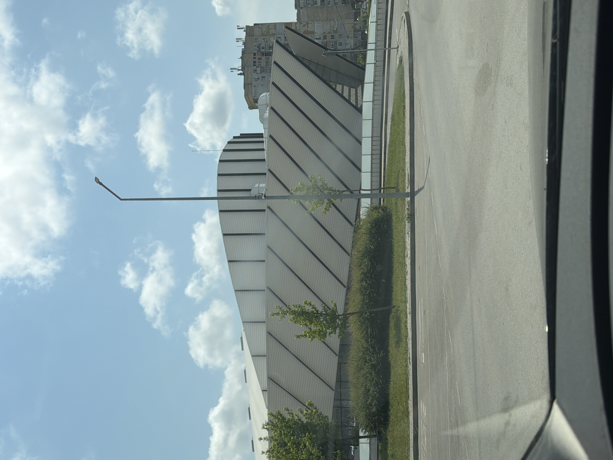 The image displays an urban daytime scene featuring contrasting architectural styles, likely taken from inside a vehicle as indicated by parts of a car frame on the right.
The central subject is a large, modern building with a distinctive design. Its facade is composed of light-colored panels separated by dark horizontal and diagonal lines, giving it a segmented, striped appearance. The building has curved elements, particularly visible on its left side. In the mid-ground, a tall, slender streetlight pole stands in front of the building.
Adjacent to the modern building is a paved road or street, which appears light gray and stretches towards the background on the right. A strip of green grass and dense bushes lines the edge of the road, running parallel to the building. Several small trees with vibrant green leaves are scattered within this green belt and in front of the building.
In the background, behind the contemporary structure, an older, multi-story residential building with numerous windows and a weathered facade is visible, offering a strong architectural contrast.
The sky above is bright blue and adorned with scattered white, fluffy cumulus clouds. The overall lighting suggests it is daytime, possibly late morning or early afternoon, with clear weather. The setting combines modern development with older urban elements. No visible text is present in the image. Given the context, this scene is located in Shumen, Bulgaria.