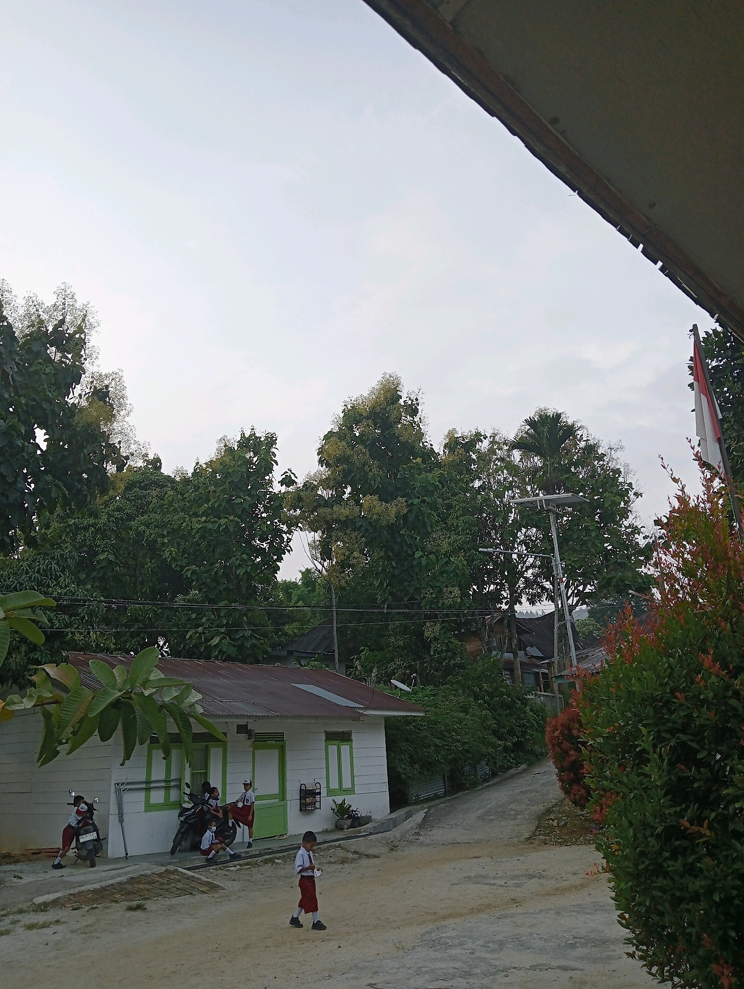 In Balige, Indonesia, a schoolyard scene unfolds under an overcast sky. A small, white building with green trim and a corrugated metal roof stands to the left, partially obscured by foliage. In front of the building, several students in uniform are gathered near motorcycles. One student walks away from the building towards the foreground, holding what appears to be paper. The ground is a mix of dirt and paved surfaces. Lush green trees with some flowering branches dominate the background, with a pole supporting electrical wires and a street light visible among them. A flagpole with a partially visible red and white Indonesian flag stands on the right. The overall atmosphere suggests a typical school day in a tropical environment.