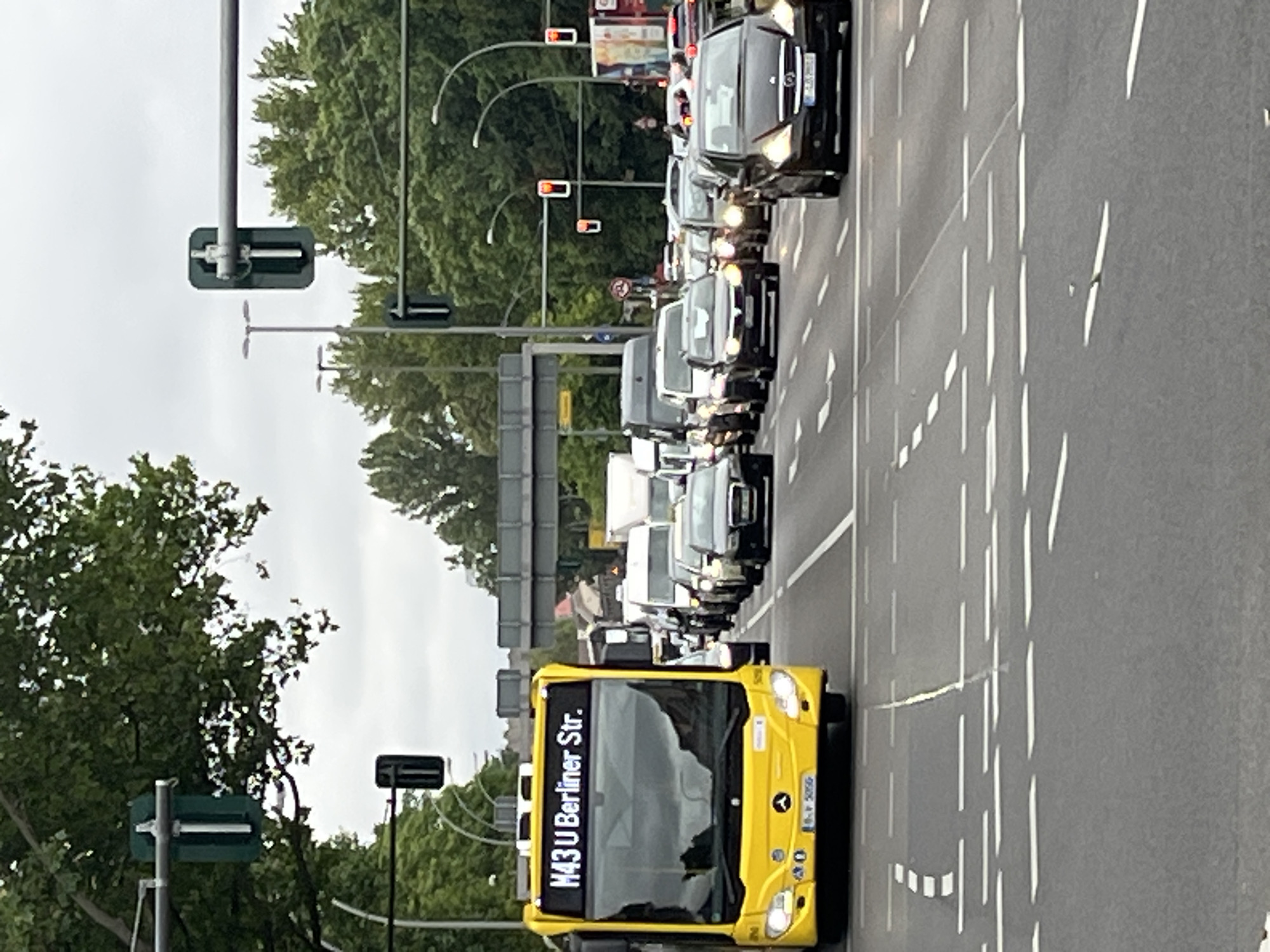 The image shows a street scene in Berlin, Germany, with a bright yellow bus in the foreground. The bus has the route number "M43 U Berliner Str." displayed on its front. The bus is in a traffic lane, stopped or moving slowly, as are the numerous cars behind it, indicating traffic congestion. Several traffic lights are visible, some with red lights illuminated, suggesting the vehicles are at an intersection. The environment is urban, with trees lining the street and a cloudy sky overhead. There are no visible pedestrians in the image. The focus is on the traffic and the yellow bus.