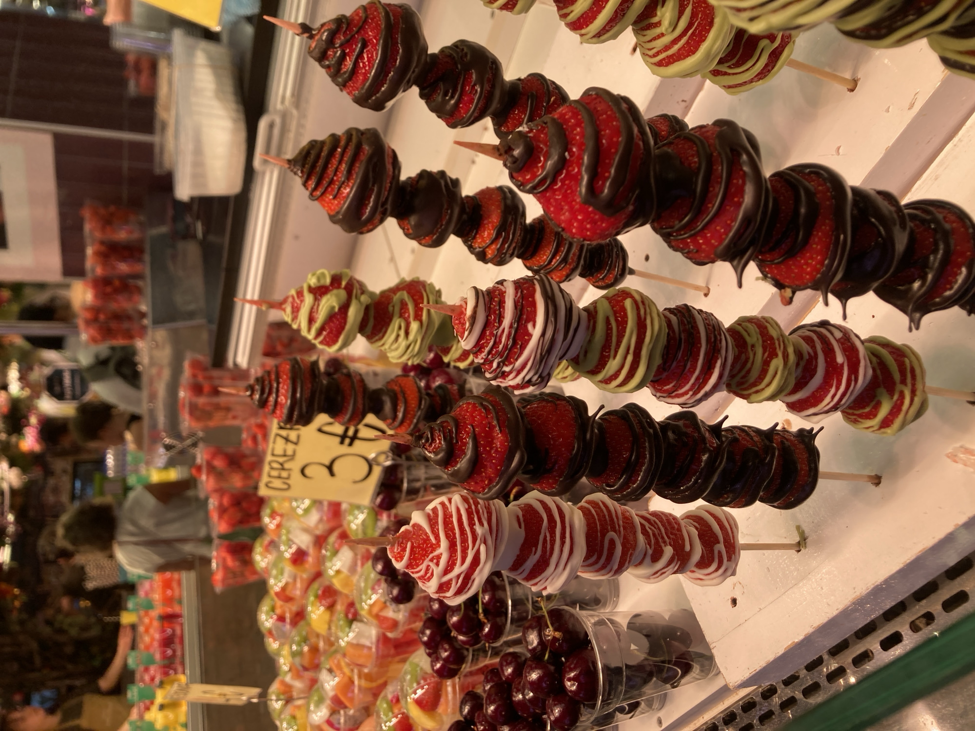 The image displays a close-up view of several skewers of fruit, primarily strawberries dipped in various types of chocolate or frosting, arranged on a white surface. Some skewers feature strawberries coated in dark chocolate, while others are drizzled with white frosting or a light green colored frosting. In the foreground, there are clear plastic cups filled with cherries and containers of cut fruit.
Behind the fruit skewers, there are glimpses of a market or food stall environment. Blurred figures of people are visible in the background, suggesting a public place. There are also stacks of packaged fruit and other food items on shelves. One skewer has a yellow price tag attached, with some visible text. The visible text on the tag reads "CEREZ...", likely indicating "Cerezas" (cherries in Spanish). The price displayed is "3 €".
The lighting suggests an indoor setting, possibly a market, during daytime. The overall scene conveys a sense of vibrant food presentation, common in bustling markets. The focus on colorful fruits and sweet coatings suggests a commercial food vendor, likely in Barcelona, Spain, given the context. The emotions evoked are primarily related to appetite and the visual appeal of fresh produce and treats.