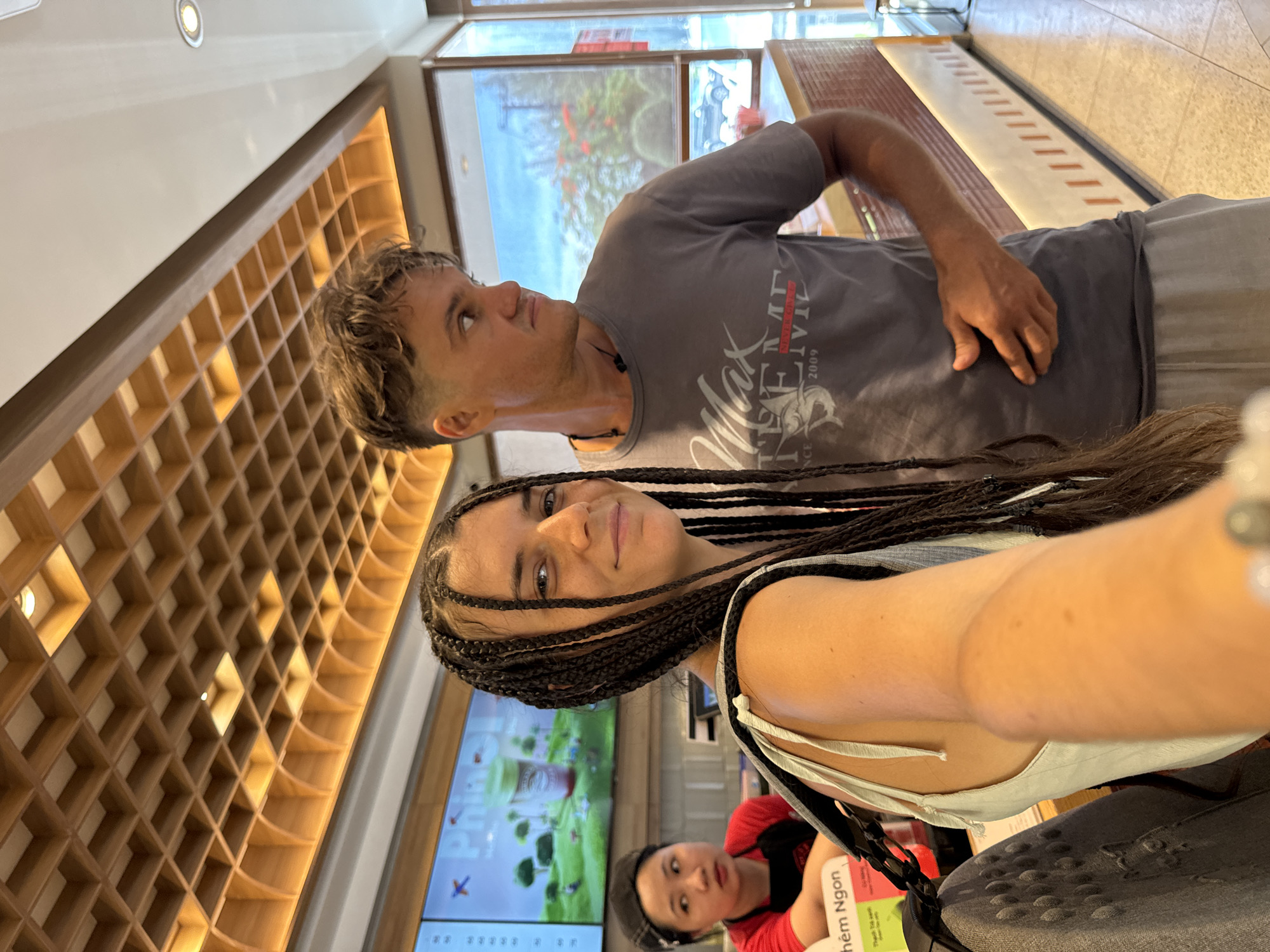 This media file shows a selfie taken by a woman with dark braided hair. She is in the foreground, looking towards the camera with a slight smile. Behind her, a man with short, wavy brown hair is looking off to the side. He is wearing a grey t-shirt with white text that appears to be a logo.
The background suggests an indoor setting, possibly a shop or restaurant, with a wall of wooden cubbies to the left. To the right, there's a window with a view of an outdoor scene, and further back, a display screen shows images of drinks and greenery. Another woman in a red shirt is visible in the background, further to the right. The overall lighting is bright, indicating it's daytime. The image captures a candid moment, likely during a casual outing.
Visible text includes "Max TEAV" and "2009" on the man's t-shirt, and "Thêm Ngon" on a green sign in the bottom right. The location context of Phan Thiết, Vietnam is provided.