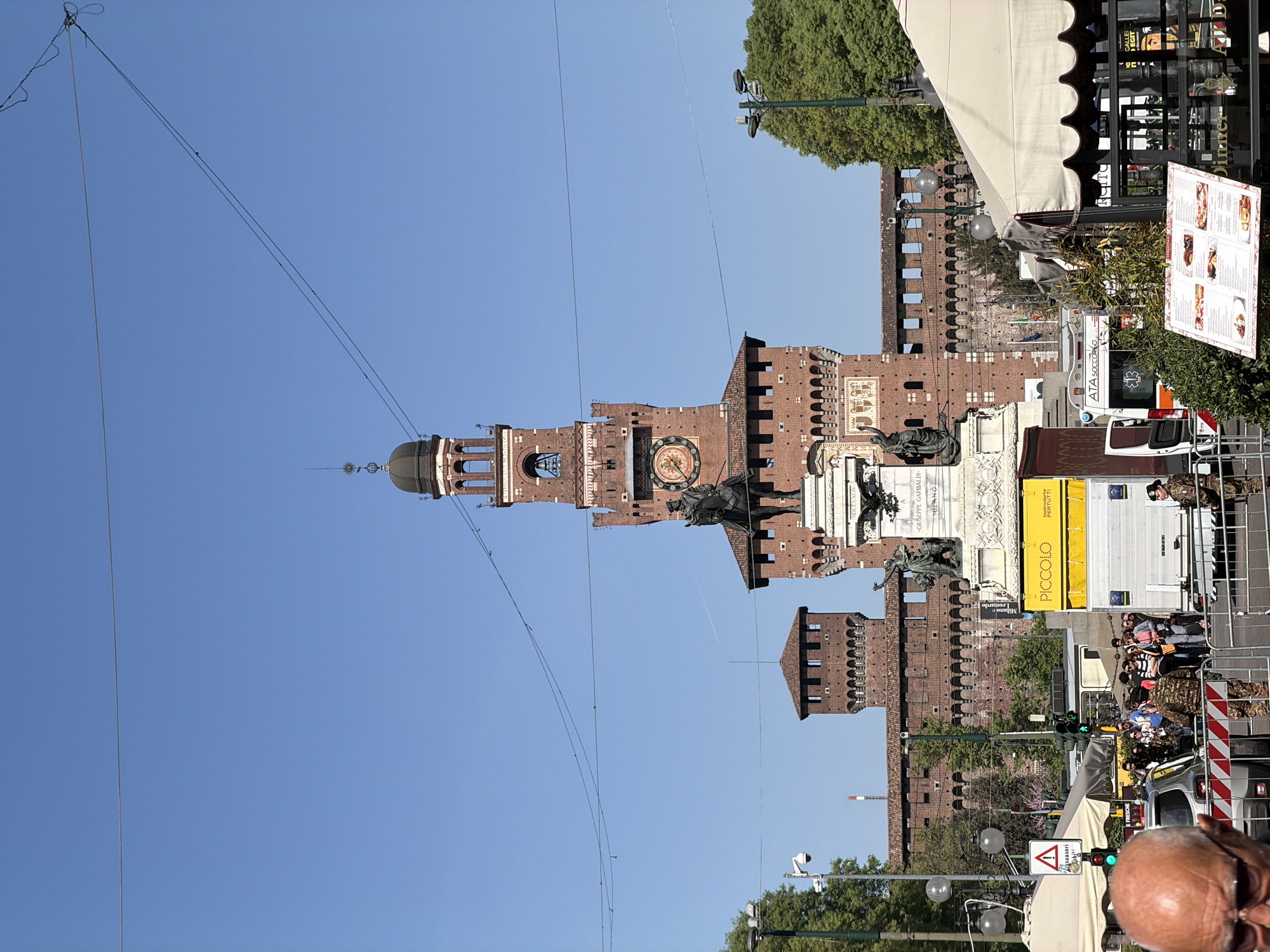 Here's a detailed description of the image:
The image depicts a view of the Sforza Castle (Castello Sforzesco) in Milan, Italy, taken from street level. The castle is the prominent feature, showcasing its brick facade, towers, and central clock tower. A statue, likely of a historical figure, stands in front of the castle. Overhead power lines extend across the upper portion of the frame. In the foreground, a busy street scene unfolds with pedestrians and service vehicles visible. Military police are present, possibly related to a public event. Street signs, restaurant awnings, and other urban elements clutter the scene. The day appears bright and sunny.