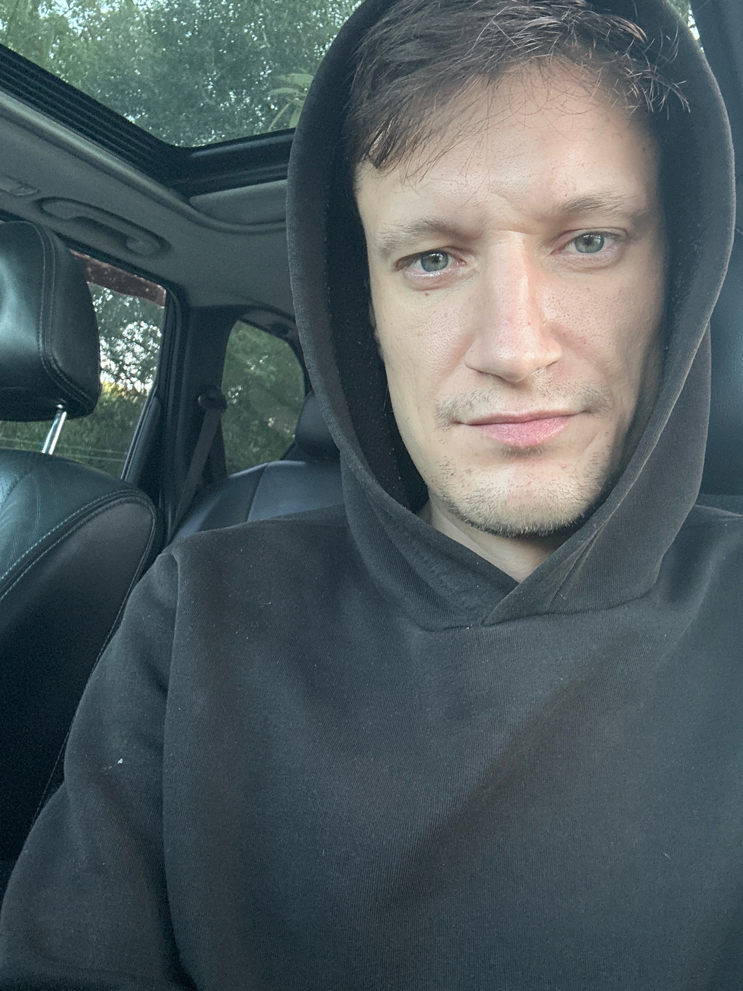 The image is a close-up, selfie-style photograph of a man in his late 20s or early 30s, seated in the driver's seat of a car. The man is wearing a dark hooded sweatshirt. His face is turned towards the camera, with his eyes looking directly into the lens. He has light skin, light brown hair, and light blue eyes. There is a faint stubble on his chin and upper lip. The interior of the car is visible behind him, including a sunroof, black leather seats, and a seatbelt. The background outside the car appears to be trees and foliage, suggesting an outdoor setting, possibly on the outskirts of a populated area. The location is identified as Makiv, Ukraine.