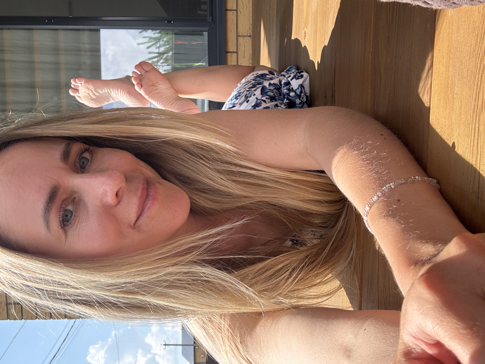 A young woman with blonde hair lies on a wooden deck outdoors, facing the camera and smiling. She is wearing a floral print dress. Her feet are crossed and visible behind her, with the soles facing upwards. The background shows a brick building with large windows and a glimpse of the sky with clouds. The scene appears to be a residential area in Chaikovsky, Russia, possibly a backyard or patio. The lighting suggests it is daytime.