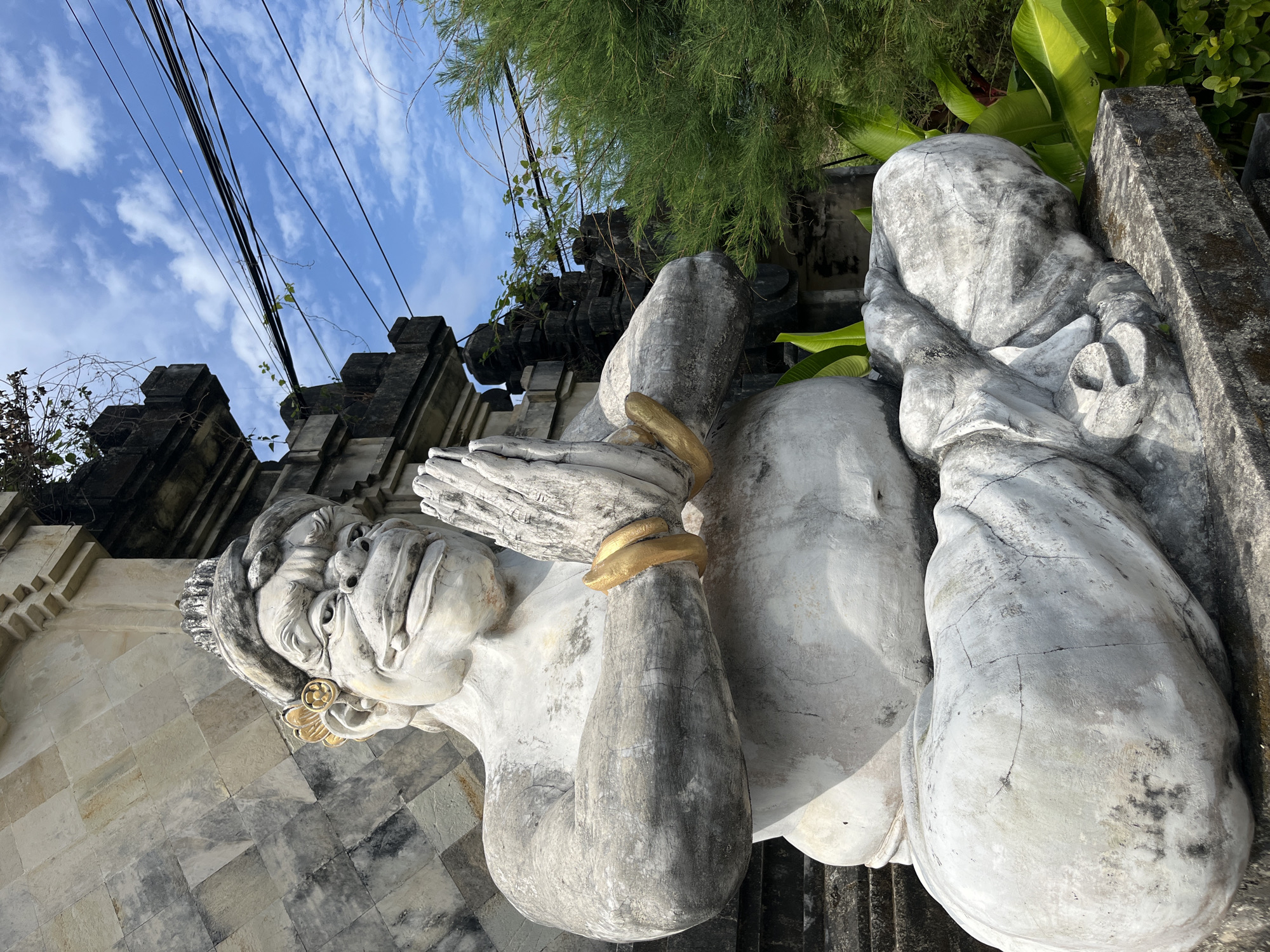 A stone statue of a large, seated figure with its hands clasped in front of its chest is shown in front of a temple-like structure. The statue is white with gray accents and has gold bands around its wrists and a gold decoration on its forehead and ear. The figure has a wide smile, prominent teeth, and large eyes. It is seated in a cross-legged position, with its legs tucked beneath it. The temple structure is made of stone blocks and has intricate carvings. Green foliage and branches are visible to the right of the statue, and electrical wires are strung across the sky in the background. The sky is blue with scattered white clouds. The statue is weathered and cracked in places. The location is identified as Kuta Selatan, Indonesia.