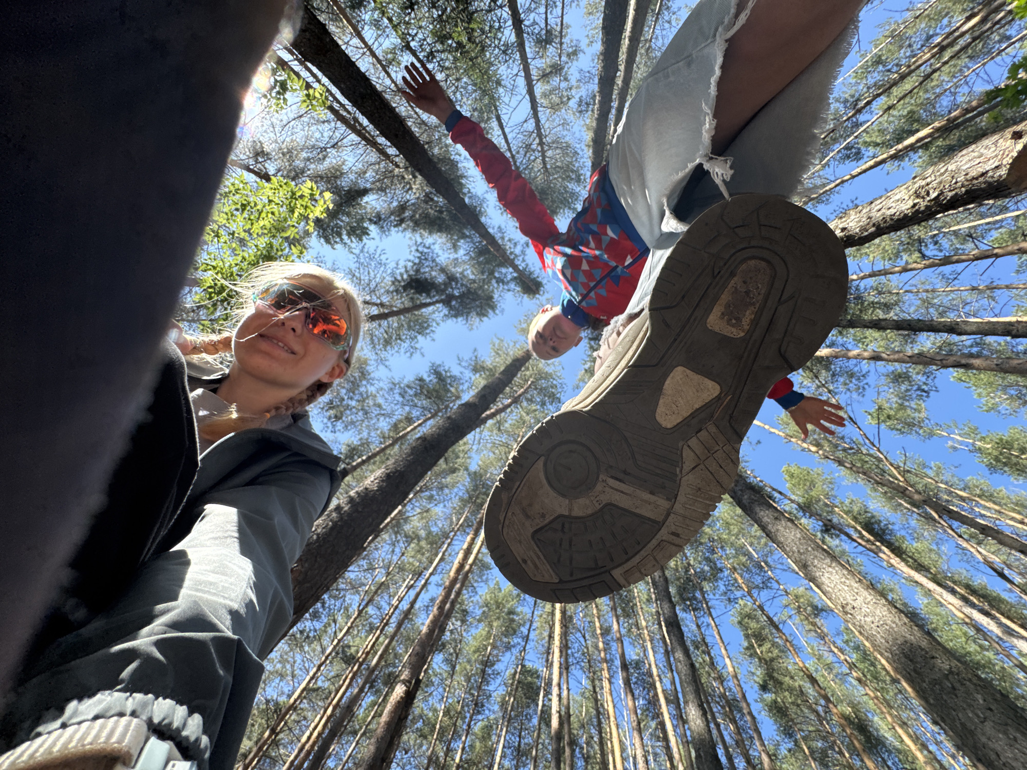 The image shows a low-angle shot of two individuals in a forest environment, with a clear blue sky and tall trees visible. The foreground is dominated by the sole of a dark-colored shoe, positioned as if someone is stepping over the camera.
In the middle ground, a person wearing a red, blue, and white patterned jacket and ripped denim shorts is seen with their arm extended, hand open, reaching towards the camera, possibly waving or interacting with the photographer.
Further back, another person is visible, wearing sunglasses with orange lenses and a grey jacket. They are smiling and looking towards the camera. Their blonde hair is braided.
The environment appears to be a pine forest, with the trees stretching upwards towards the sky. There is no indication of urban infrastructure or any specific landmarks that would identify the location within Chaikovsky, Russia. The scene depicts a casual outdoor activity, likely hiking or exploring in a natural setting.