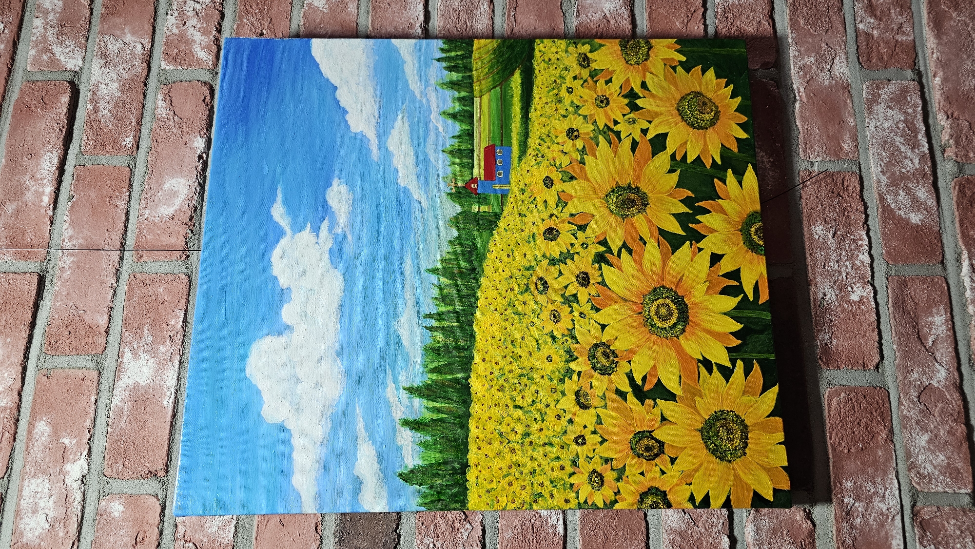 A painting of a sunflower field under a blue sky with white clouds is displayed on a brick wall. The painting depicts a vast field of sunflowers in full bloom, with their bright yellow petals and dark centers dominating the foreground and stretching towards the horizon. In the middle ground, a dense line of green trees forms a backdrop to the field, and a small blue building with a red roof and three white windows, possibly a church or a house, is situated to the right of the trees. The sky is a vibrant blue, dotted with fluffy white clouds. The painting itself is rectangular and appears to be made of canvas. It is hung on a textured brick wall, with visible mortar between the bricks. The overall scene evokes a sense of warmth and rural serenity. The artwork is located in Asan-si, South Korea.