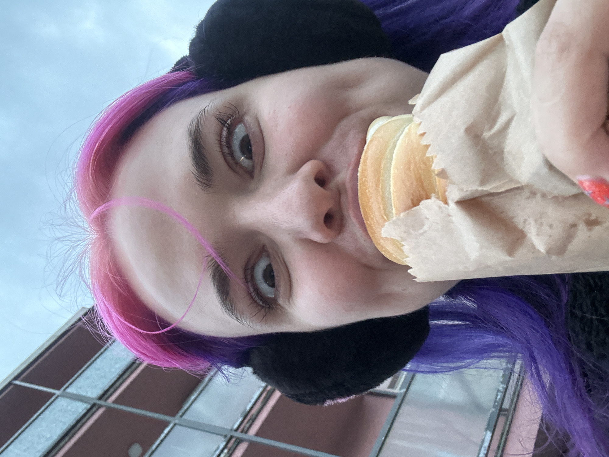 A close-up, rotated selfie features a young person with two-toned hair (pink and purple) wearing black earmuffs. The person is holding a sandwich in a brown paper bag and taking a bite. The bottom of the shot reveals an exterior building with mirrored windows. The location is identified as the city of Zagreb, Croatia.